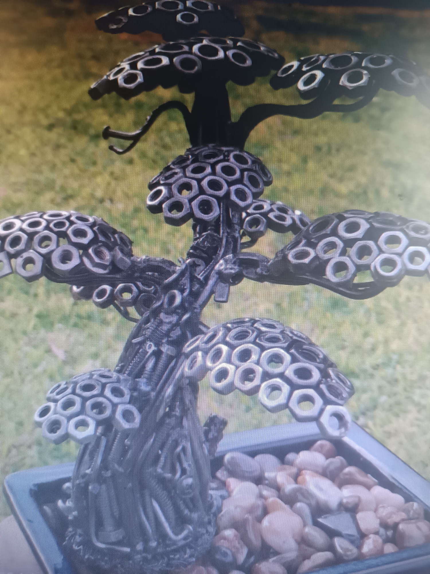 An industrial art sculpture resembling a bonsai tree, constructed from metal nuts and bolts, is displayed in a planter filled with pebbles. The sculpture is dark in color and features multiple levels of "foliage" made from interlocking nuts. The background consists of a blurred green and yellow grassy area, suggesting an outdoor setting. No people are present in the image. The location of Klyuchi, Russia is not discernible from the visual information provided.