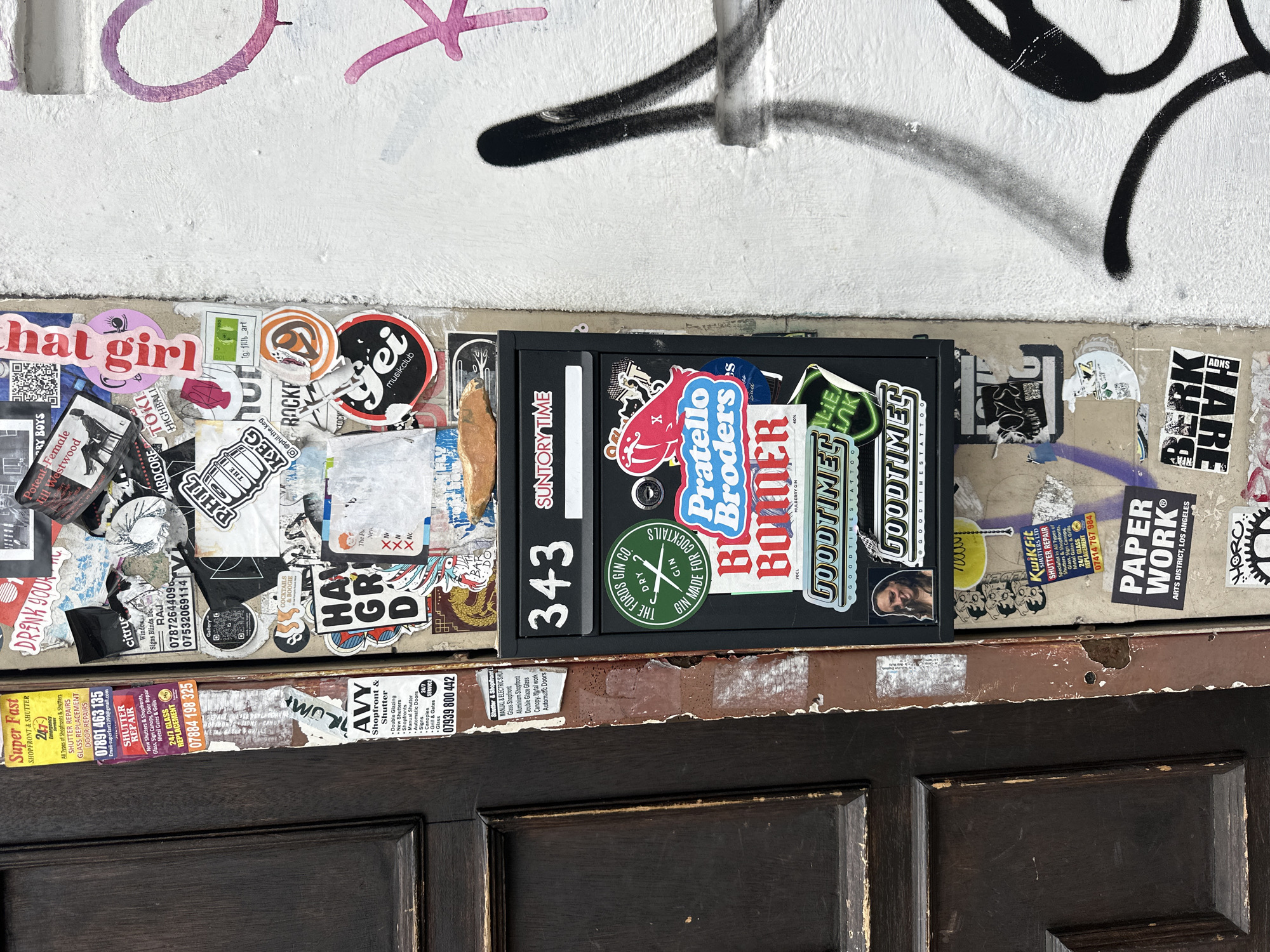 The image presents a cluttered urban scene. A black metal box, possibly a mailbox or utility box, is centrally located and adorned with numerous stickers of various sizes, colors, and designs, including stickers for "Pratello Broders," "Goodtime Tattoo," and "SuntoryTime," along with the number "343". The box is mounted on a narrow, weathered strip of wood covered in additional stickers and partially obscured by the box itself. Below the wooden strip is a dark brown, paneled door, presumably of a building. Above the box, the wall is graffitied with pink and black markings on a white painted surface. The scene is further cluttered with stickers, flyers, and advertisements plastered across the surfaces to the left and right of the box, with visible texts like "Super Fast Shopfront & Shutter," "Shutter Repair," and "Avy Shopfront & Shutter". The setting appears to be an exterior wall of a building in an urban environment. While the exact location in London, United Kingdom, is not specified, the presence of local business advertisements and graffiti suggests a vibrant, densely populated area.