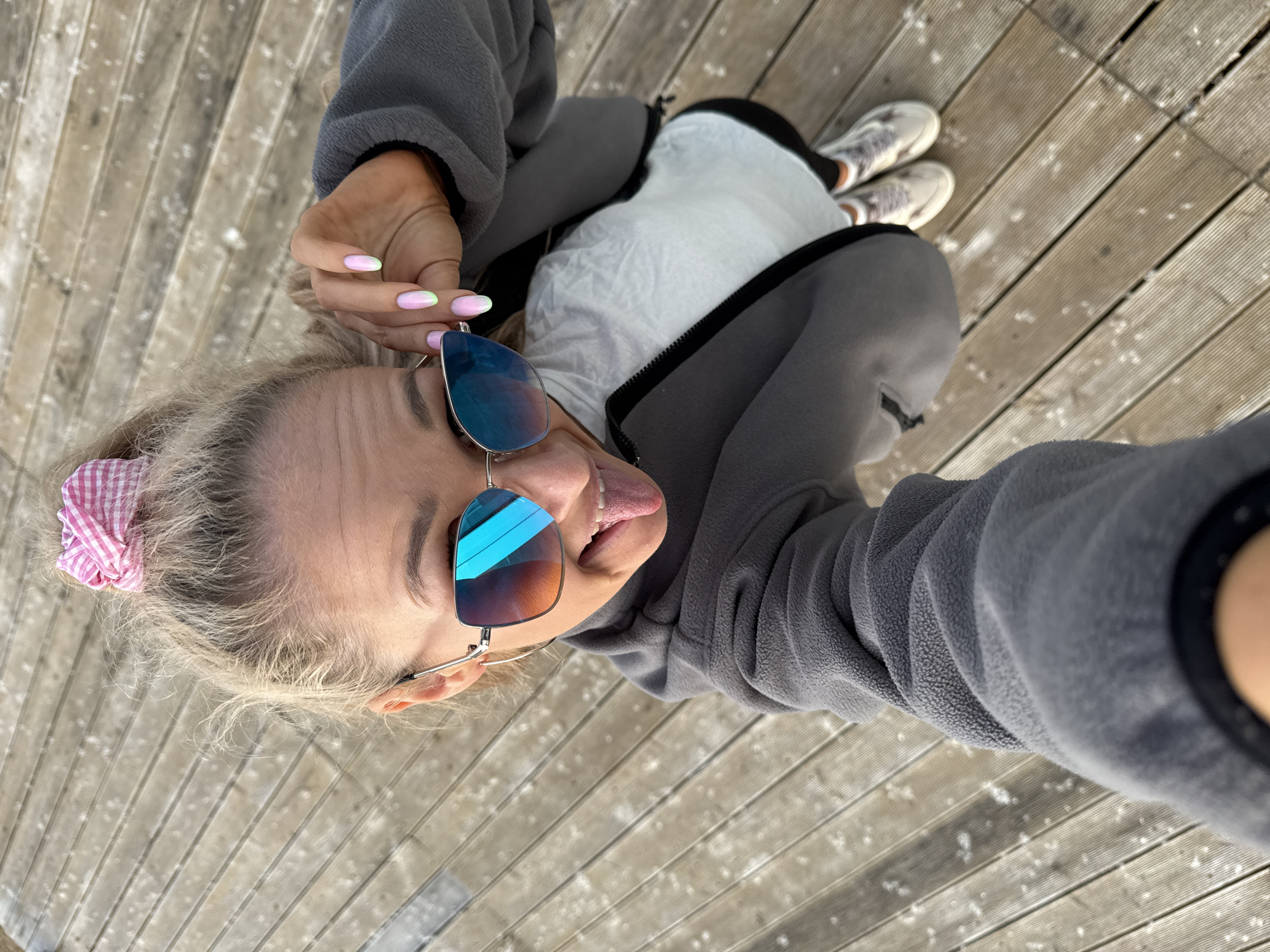 A high-angle selfie captures a blonde female, approximately 20-35 years old, playfully sticking her tongue out. She wears large, rectangular, silver-framed sunglasses with blue and orange reflective lenses, which she is adjusting with her right hand. Her fingernails feature an ombre design, transitioning from light pink to light green. Her blonde hair is tied back in a messy bun secured with a pink and white checkered scrunchie. She is dressed in a grey fleece zippered jacket over a white top and black leggings. Light-colored athletic shoes with a subtle pattern are visible on her feet. The subject is standing on a light-toned wooden surface, possibly a deck or boardwalk, composed of parallel planks. The wooden surface is speckled with numerous small, white, irregular particles, consistent with light snow or frost. The scene takes place outdoors in the city of Chaikovsky, Russia, under bright daylight conditions.
