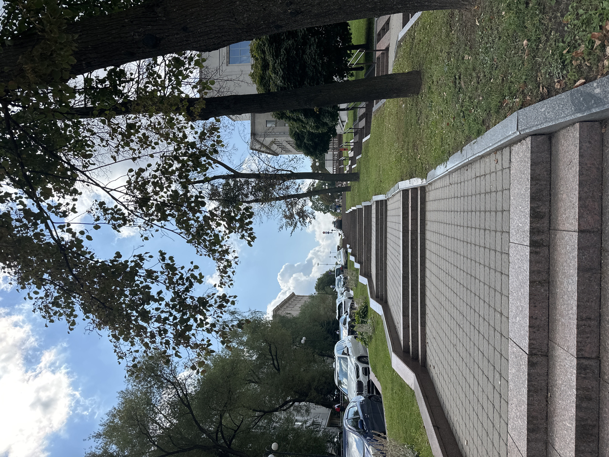 The image captures an outdoor perspective of a grand staircase with granite steps and a paved walkway, flanked by a grassy slope and mature trees. The staircase ascends towards the upper reaches of the image, leading to buildings in the background. To the left, several cars are parked along the street, suggesting an urban setting. The sky above is a mix of blue and white clouds. The scene appears to be situated in Minsk, Belarus, potentially in a park or a public area with notable architecture. The overall impression is one of a well-maintained public space on a bright day.