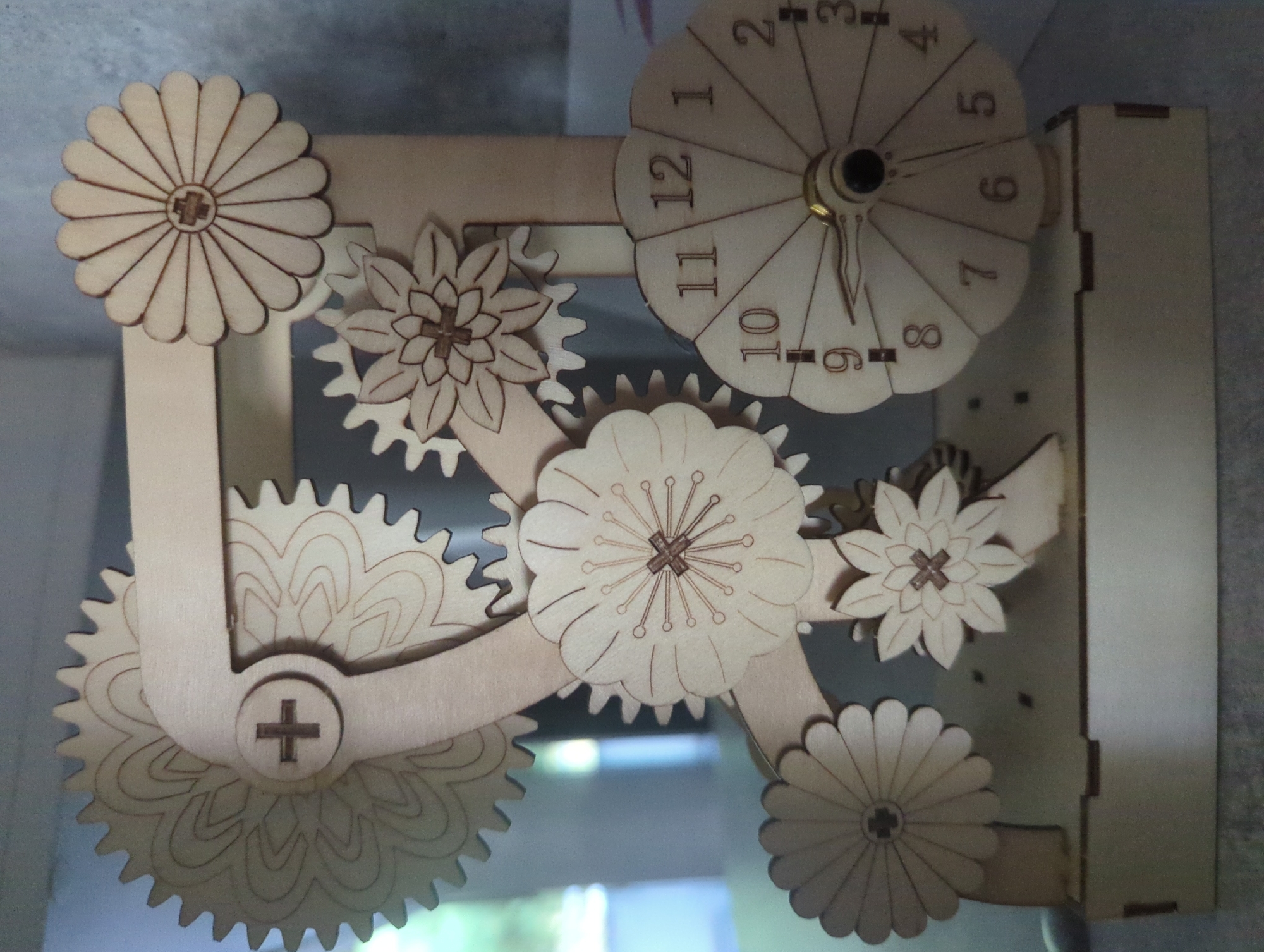 The image shows a wooden mechanical clock with a decorative gear system. The clock face has numbers from 1 to 12, and hands indicating the time. The clock is adorned with wooden gears and floral designs, giving it an artistic and intricate appearance. The background appears to be a neutral, indoor setting, possibly a workshop or display area. There are no people or animals visible in the image. The scene depicts a stationary object. The image does not provide any information that allows for the determination of its location in the city of Iserlohn, Germany.