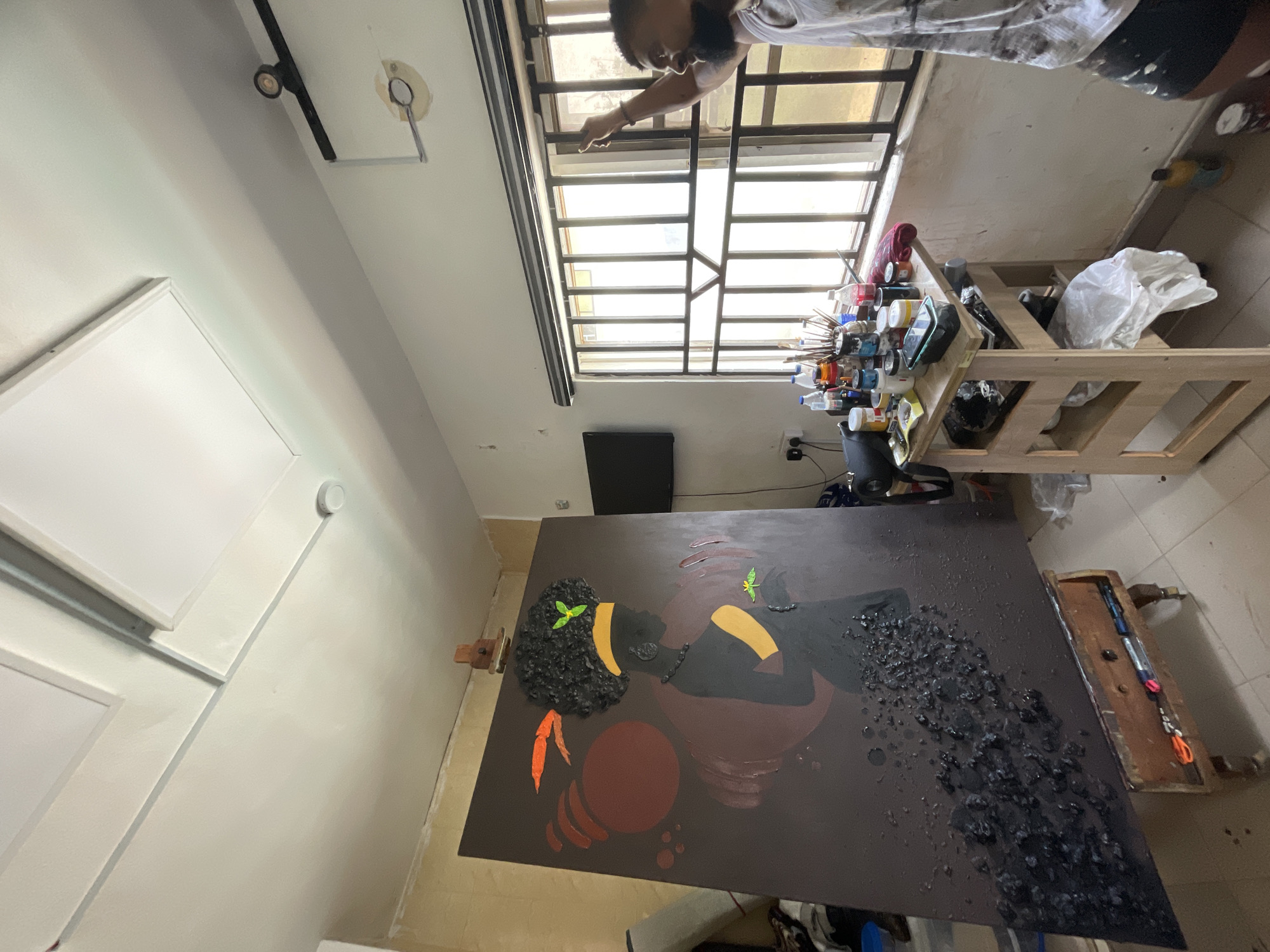 An indoor scene shows a partially completed artwork depicting a Black woman with textured hair and a red sphere positioned in the lower left corner of the frame. The painting rests on an easel. A person, possibly the artist, stands near a window with barred panes. To the right of the window, a wooden shelf holds assorted art supplies. The room appears to be a studio with white walls and tile flooring. The location is likely in Ado, Nigeria, based on contextual information not directly visible in the image but implied by the prompt.