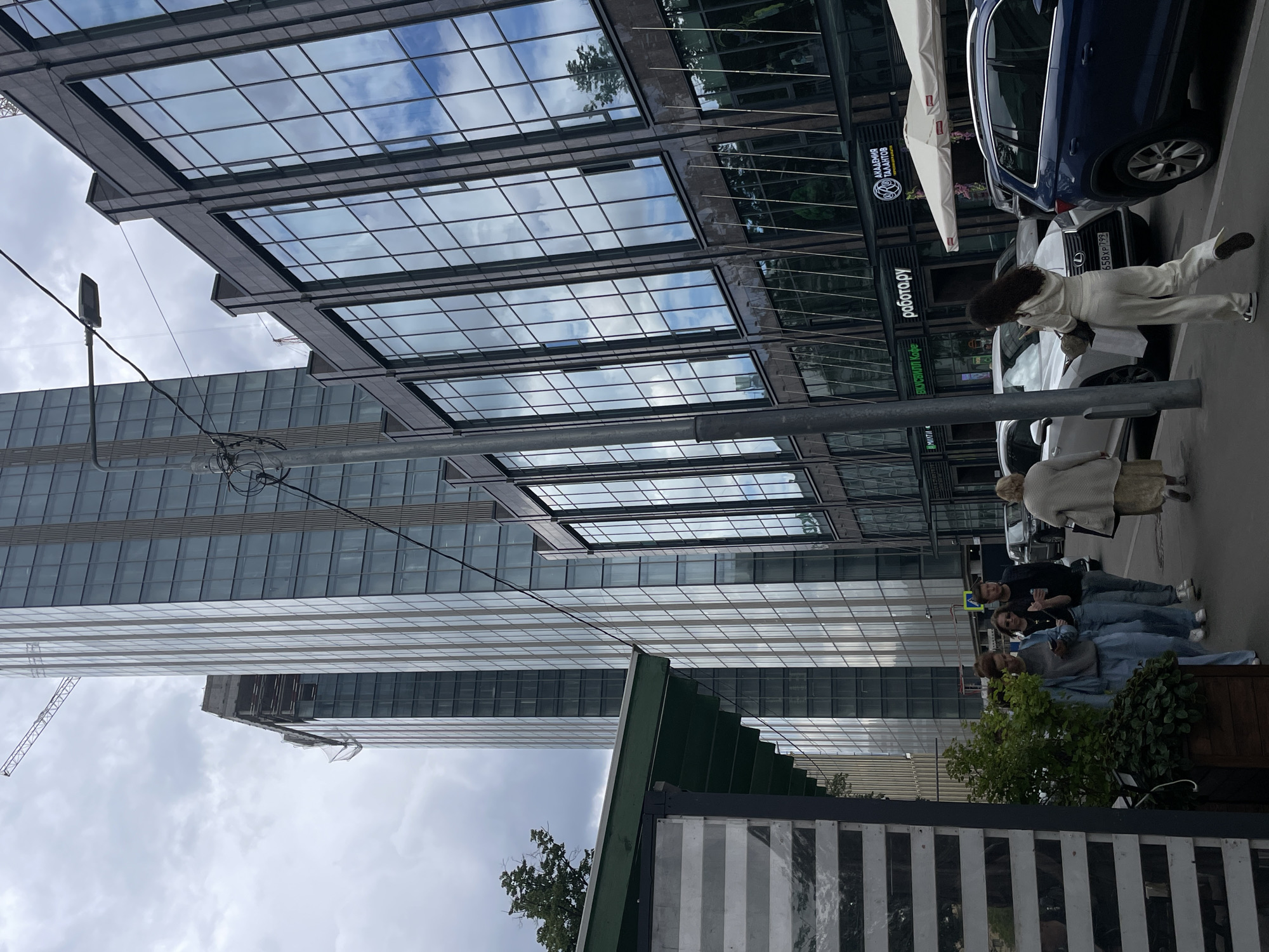 The image captures a street scene in Moscow, Russia, characterized by modern architecture and overcast skies. Dominating the background are two tall, glass-fronted skyscrapers, one appearing to be under construction with exposed structural elements and netting. In the foreground and midground, a lower, similarly modern building with large glass windows stretches along the street. These windows reflect the sky and surrounding environment.
A street lamp on a pole is visible in the middle ground. On the sidewalk, several individuals are walking. One woman in a light blue tracksuit is looking at her phone. Behind her, a man and a woman, both in casual clothing, are walking. Further down the street, another woman in a beige cardigan and dress is walking away from the camera, carrying a bag.
Several cars are parked along the street, including a blue SUV and a white sedan. A white tent or awning is set up outside a building, suggesting a cafe or outdoor seating area. Signs on the buildings indicate businesses, with legible text including "работа.ру" and "ВКУСВИЛЛ Кафе". To the left, a portion of a structure with horizontal striped panels and a green roof is visible. Trees and greenery are present, adding a touch of nature to the urban landscape. The overall impression is a bustling city street with a mix of contemporary commercial and residential buildings.