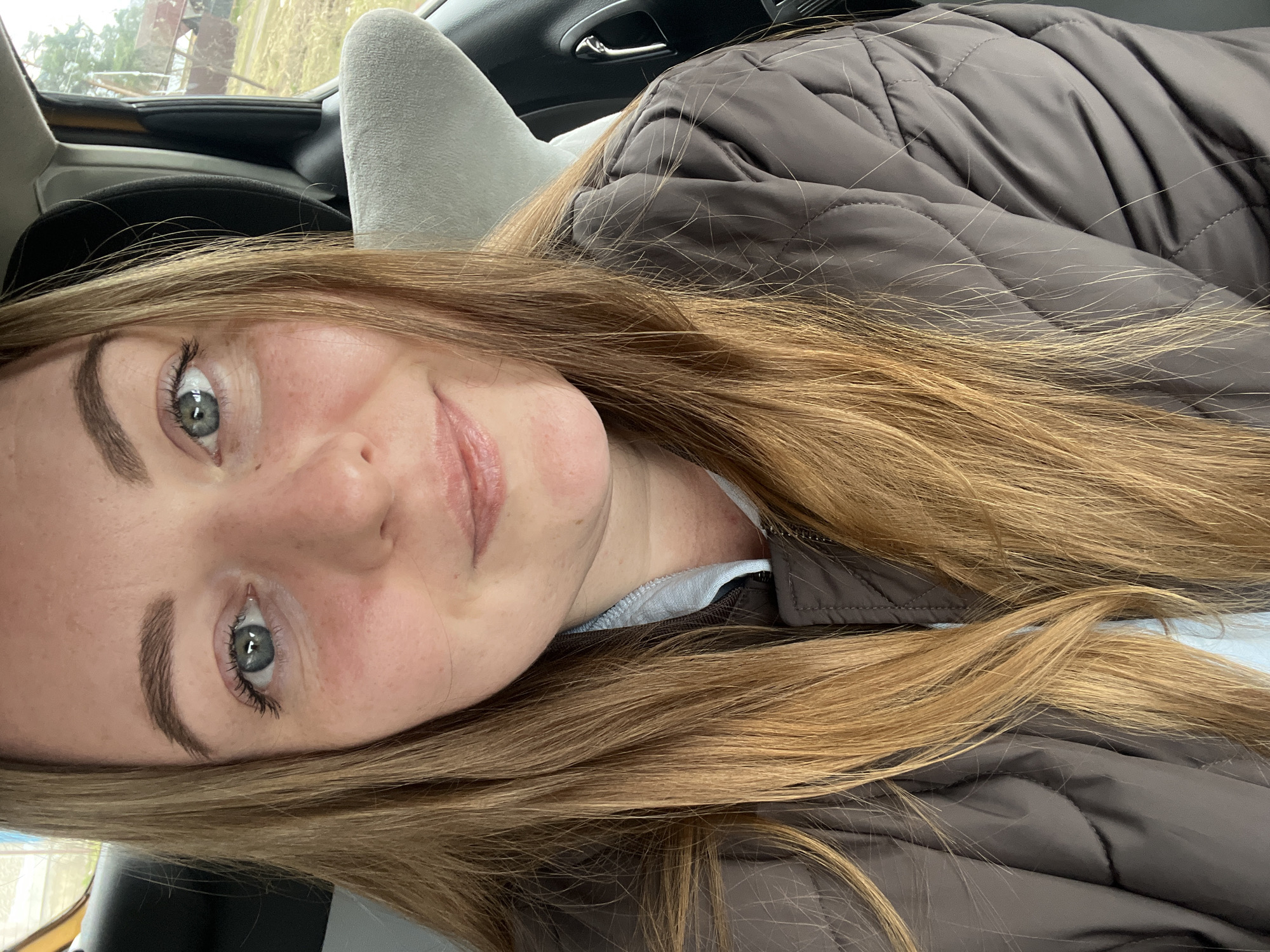 A fair-skinned woman with light brown hair and gray eyes is depicted in a close-up, slightly angled portrait. She appears to be indoors, possibly seated in a vehicle, as indicated by the headrest and car interior visible in the background. She is wearing a brown quilted jacket over a light-colored top. The background shows blurred details of the car interior and a glimpse of an outdoor scene, with trees visible through the window. There is no clear indication to confirm that it is in the city of Раздолье, Russia.