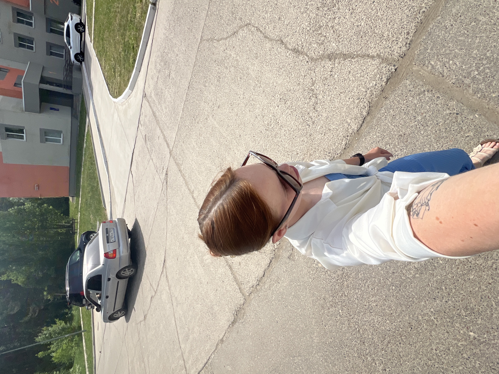 A medium-shot, high-angle photograph shows a person on a light gray concrete surface. The individual is wearing sunglasses, a white blouse, and blue pants, with beige sandals on their feet. Their hair is reddish-brown. A tattoo is visible on their upper arm. The background features apartment buildings, cars, and green vegetation, suggesting an urban environment. The location is Chaikovsky, Russia.
