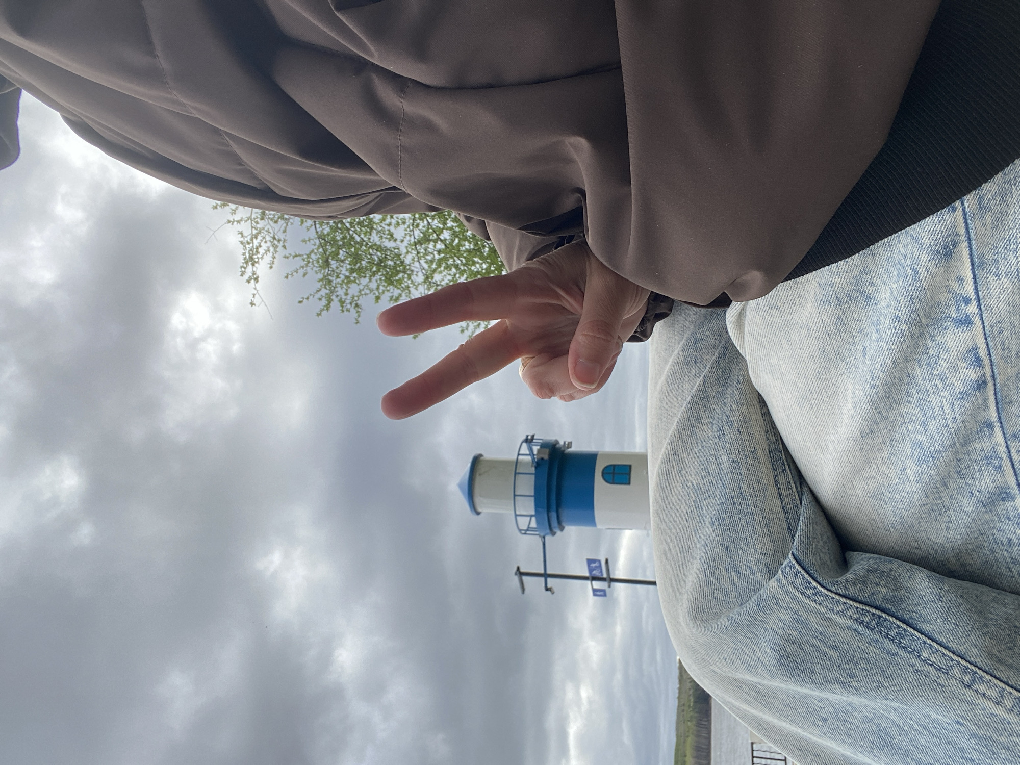A low-angle shot looking upward, featuring a person in a brown jacket and light blue jeans, giving a peace sign. Behind them is a small, blue-and-white lighthouse replica against a cloudy sky. Based on the lighthouse design, this is likely located in the city of Chaikovsky, Perm Krai, Russia.