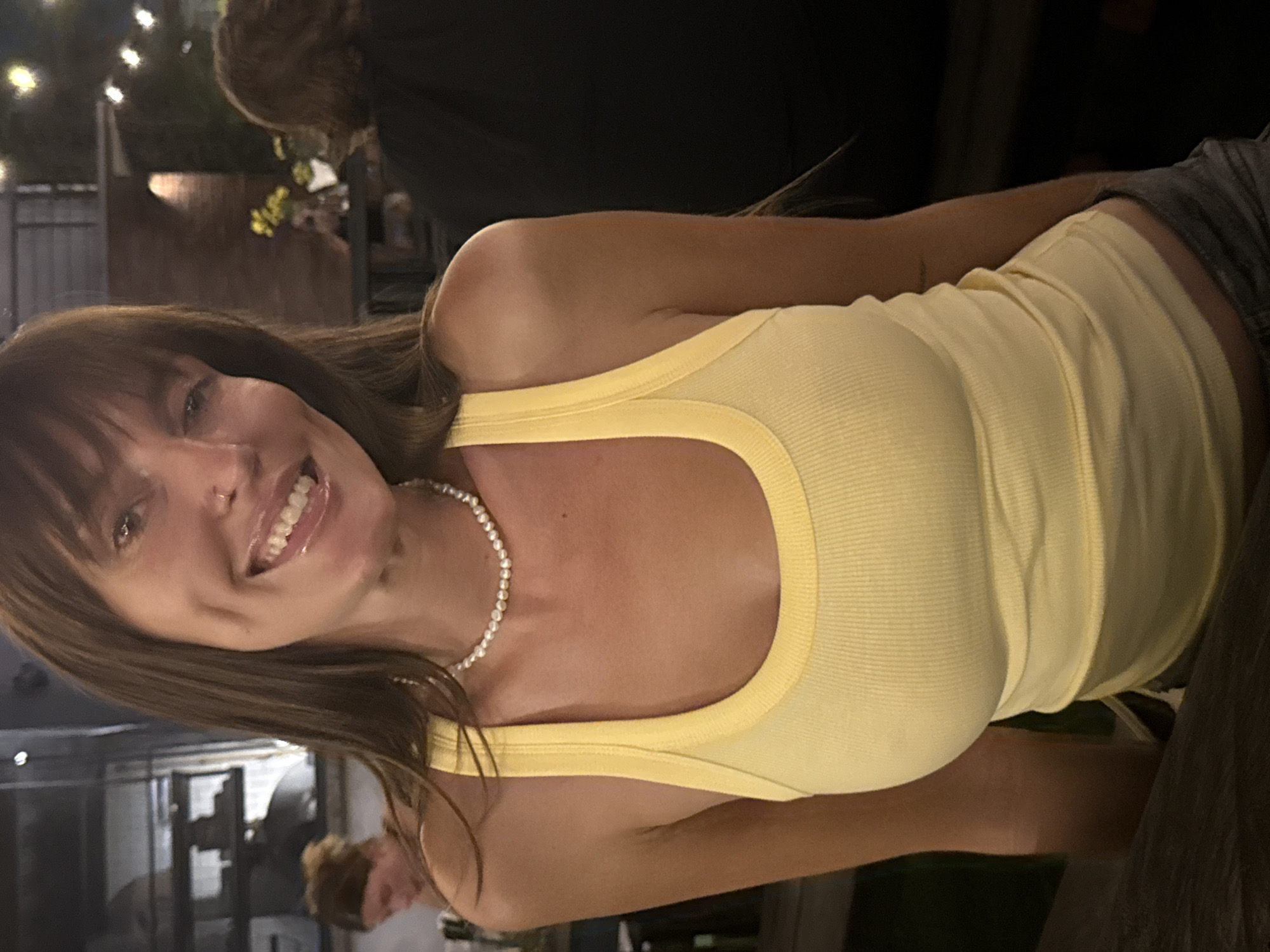 A close-up, portrait-oriented photograph, rotated 90 degrees counter-clockwise, depicts a woman smiling brightly at the camera in a dim, evening setting, likely in North Kuta, Indonesia. She occupies the left and central portion of the frame.
The woman has long, straight brown hair with bangs framing her forehead. She is fair-skinned with a tanned complexion and displays a wide, cheerful smile that shows her teeth. Around her neck, she wears a pearl necklace. Her attire consists of a light yellow, ribbed tank top with a contrasting yellow trim along the neckline and straps.
The background is softly blurred and dimly lit, featuring indistinct string lights in the upper portion, suggesting an outdoor or open-air venue. The backs of other patrons are partially visible; one person wearing a dark top is seen in the upper right, and another with reddish-brown hair is in the lower-left. The low light levels confirm an evening or nighttime event. The woman's joyful expression indicates a pleasant and social atmosphere. No text is visible in the image.