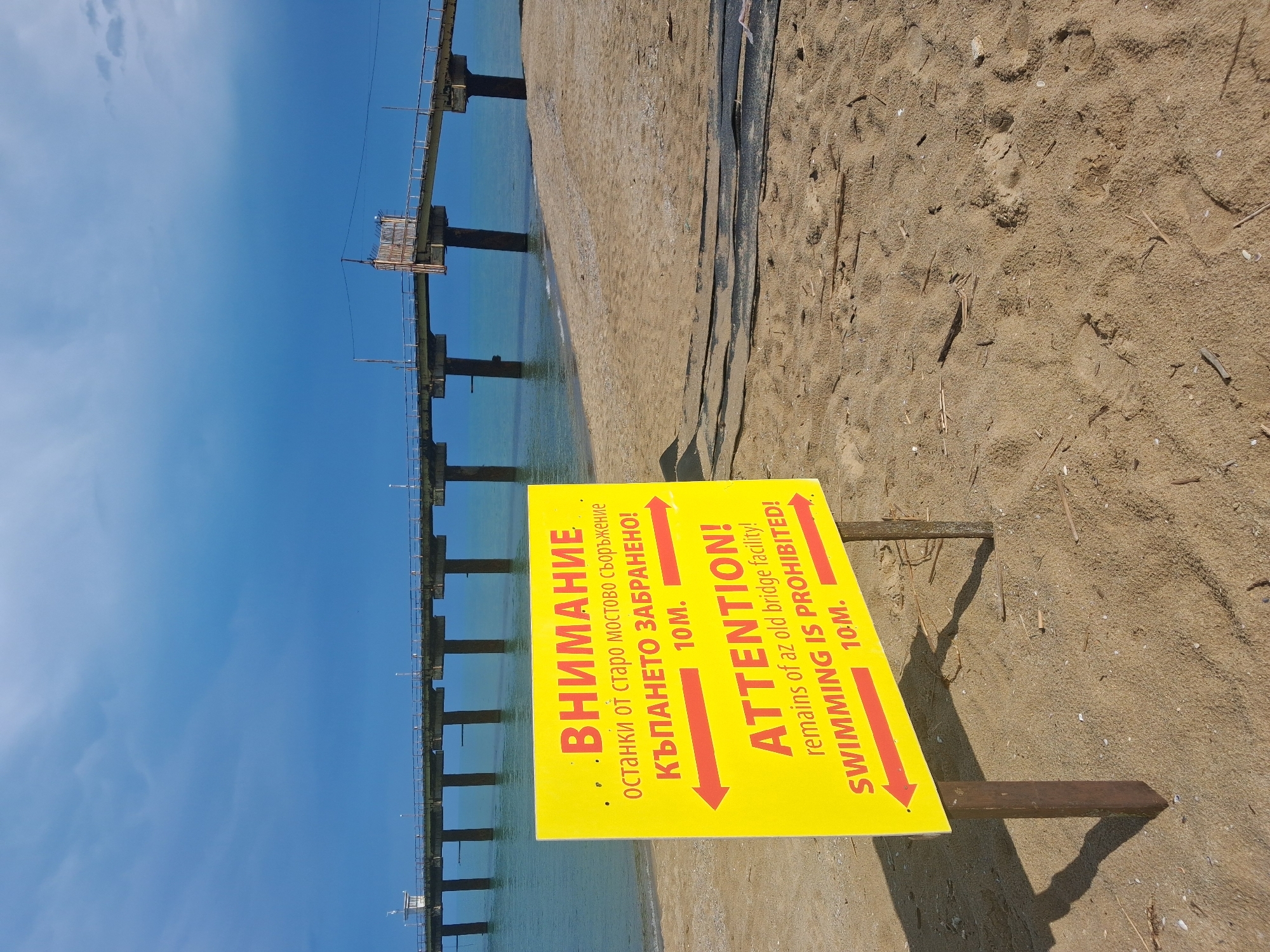 This image depicts a coastal scene in Shkorpilovtsi, Bulgaria, featuring a beach, the sea, and a concrete pier under a dramatic, overcast blue sky. In the foreground, a large yellow warning sign is centrally positioned on two brown wooden posts, casting distinct shadows on the sand.
The sign displays text in both Bulgarian (Cyrillic script) and English. The Bulgarian text reads "ВНИМАНИЕ останки от старо мостово съоръжение КЪПАНЕТО ЗАБРАНЕНО! 10М.", translating to "ATTENTION! remains of an old bridge facility SWIMMING IS PROHIBITED! 10M.". The English text below it confirms "ATTENTION! remains of az old bridge facility! SWIMMING IS PROHIBITED! 10M!". Two prominent red arrows point left and right on the sign, indicating the prohibited area's lateral extent from the pier.
The sandy beach, a light tan color, occupies the lower portion of the frame. It shows various footprints and scattered natural debris such as small sticks and shells. Closer to the water's edge, several dark, irregular, elongated objects (possibly industrial fabric or debris) are partially buried in the sand.
The sea, a calm turquoise hue, stretches across the middle ground. A long, multi-sectioned concrete pier extends from the left side of the frame, curving slightly as it projects into the water towards the right. The pier is supported by numerous thick, dark-colored concrete pillars. A white metal railing runs along both sides of the pier's deck, which also features intermittent lampposts. On the far left of the pier, a small, white, box-like structure with a roof stands. Further along the pier to the right, a makeshift barrier constructed of vertical wooden slats or reeds obstructs the path.
The sky is a deep, dramatic blue with areas of lighter blue and visible cloud formations, suggesting a change in weather or specific lighting conditions. No people are visible in the scene.