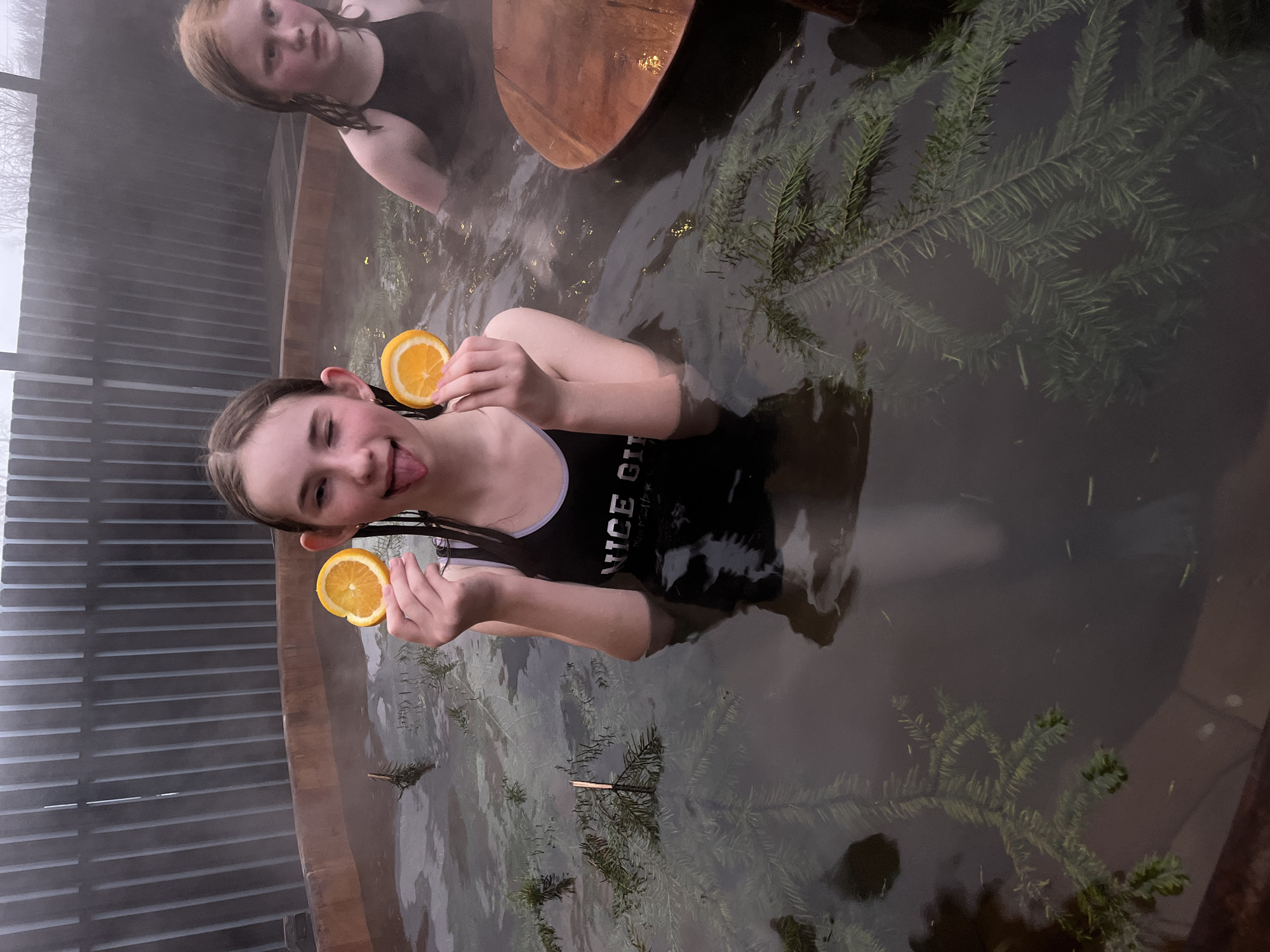 Two young females are inside a circular wooden tub containing water and evergreen branches. One female, positioned closer to the viewer, holds two orange halves near her face and sticks out her tongue. She wears a black bathing suit. The other female is in the background, partially submerged, wearing a dark top. The environment appears to be outdoors, with wooden planks visible on one side and a cloudy sky.