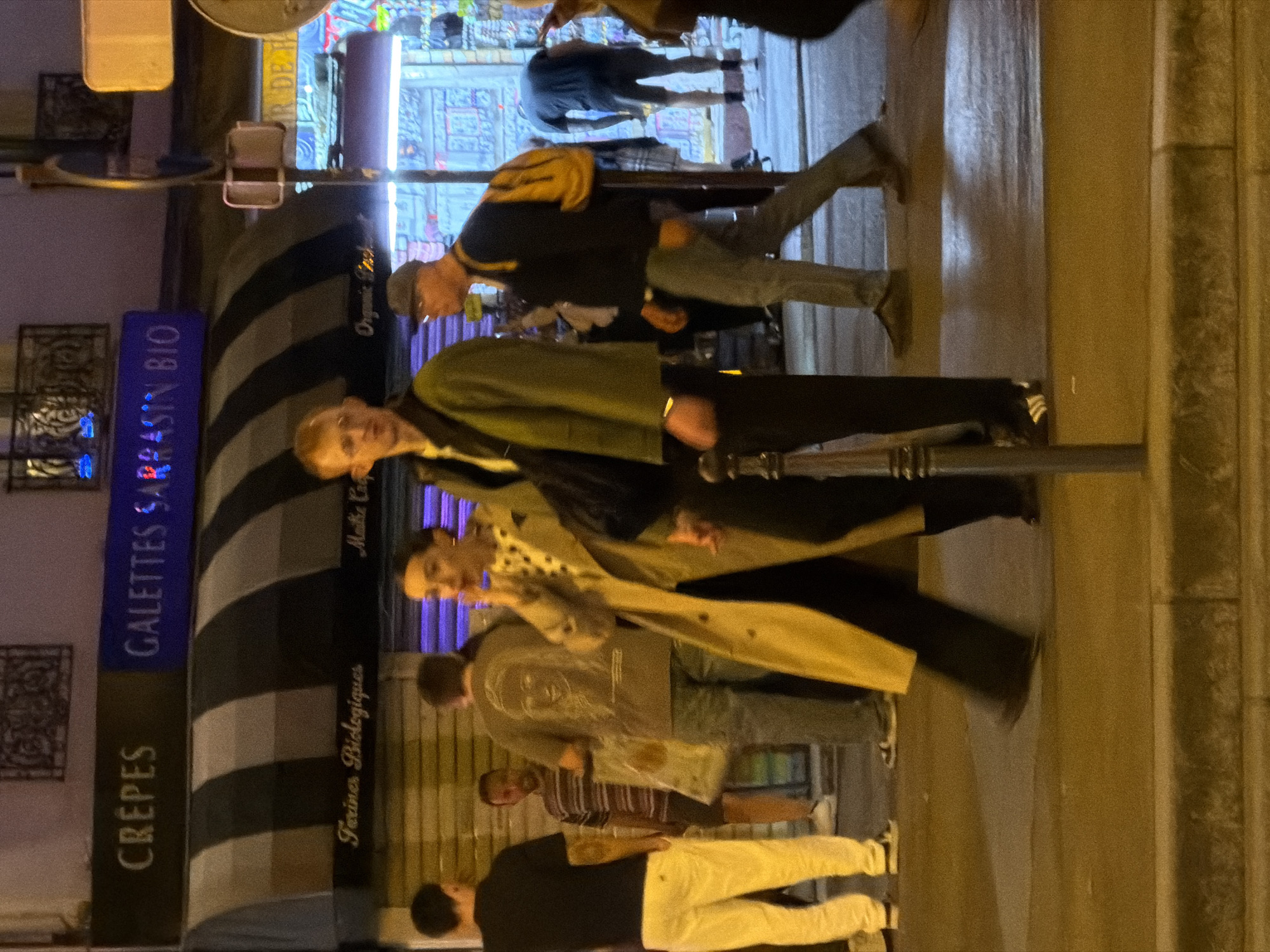 The image captures a street scene, possibly in Paris, France, at night. The illumination suggests artificial lighting, highlighting pedestrians outside a food establishment.
A sign indicates "GALETTES SARRASIN BIO CRÊPES." People are observed walking, standing, and engaging in apparent conversation. The environment includes storefronts and street infrastructure typical of an urban setting. Clothing styles vary, reflecting casual attire.
