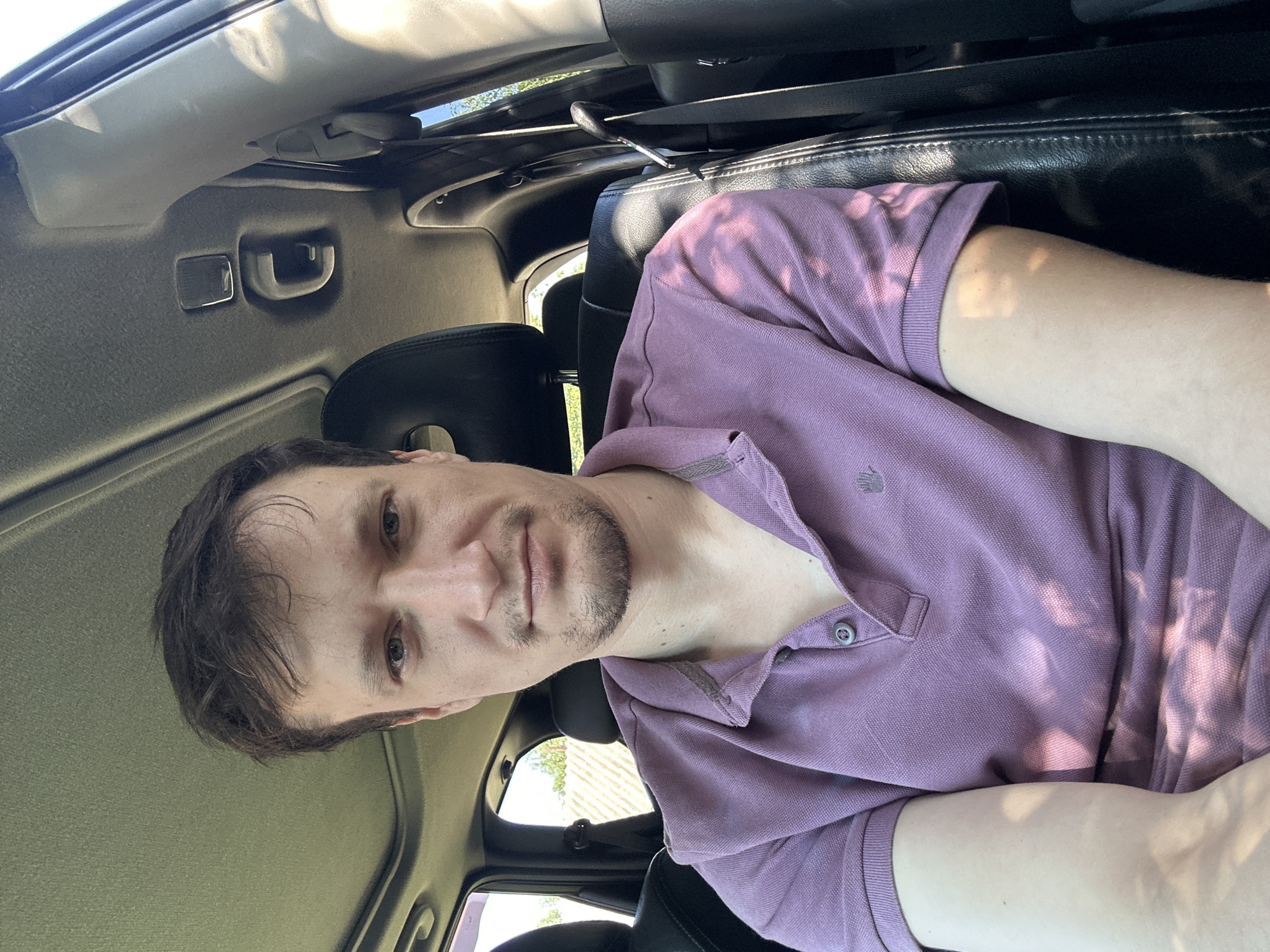 A medium shot is captured from inside a vehicle. The subject, a white male with short, dark hair and a light beard, is seated in the driver's seat, looking directly at the camera. He is wearing a light purple polo shirt with two buttons fastened at the collar. The car's interior is visible, including black leather seats, a sunroof, and a seatbelt. The background shows a fence and greenery, indicating an outdoor setting, possibly on the outskirts of a settlement. The lighting suggests it is daytime, with sunlight casting dappled shadows on the subject and interior.
