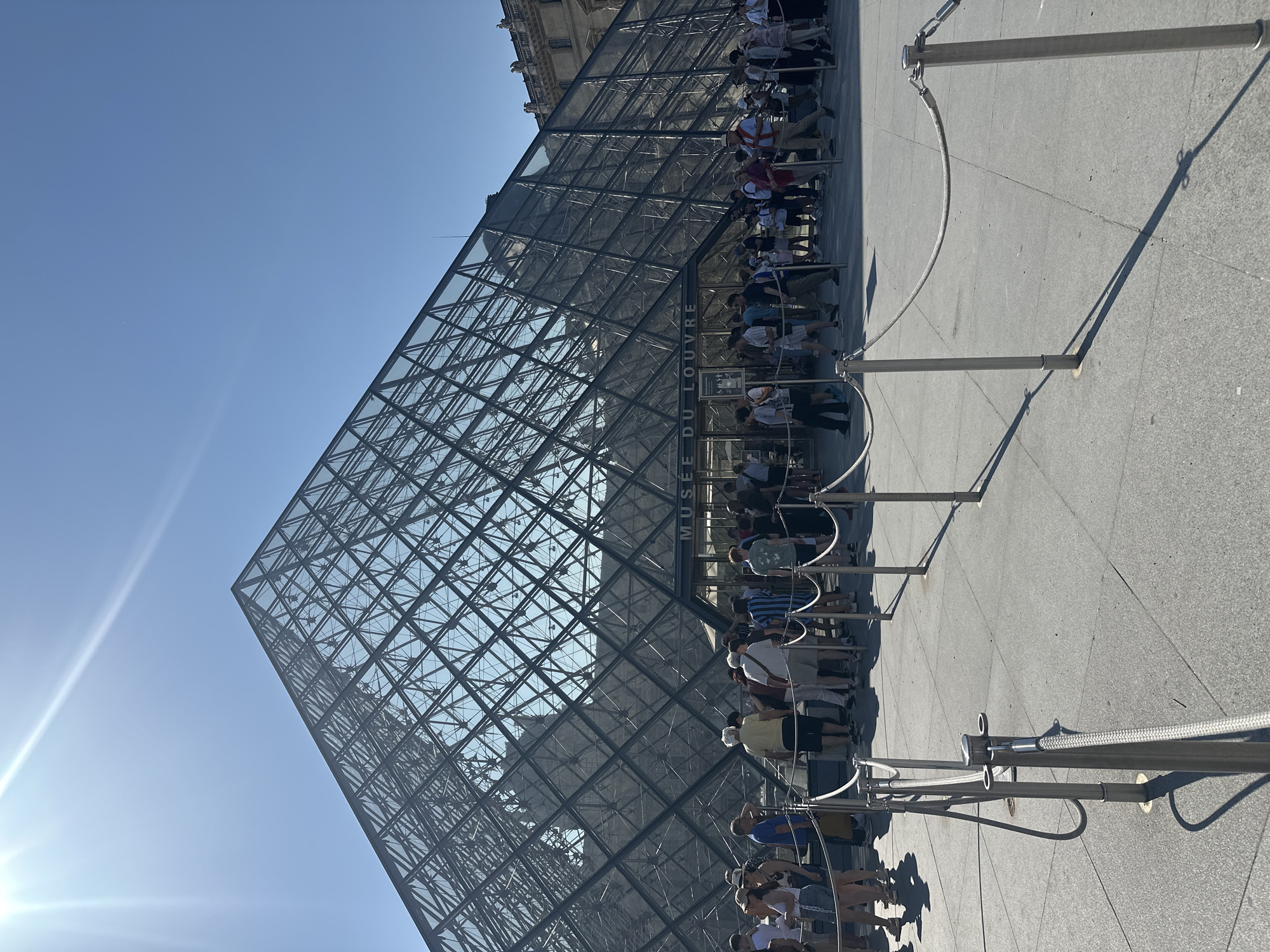 The image captures the iconic glass pyramid of the Louvre Museum in Paris, France. Under a clear, bright blue sky with a strong sun casting sharp shadows, a diverse group of people are queuing in a roped-off line leading towards the museum entrance. The pyramid's geometric glass and metal structure is prominently displayed, with the words "MUSEE DU LOUVRE" visible above the entrance doors. Visitors are dressed in casual summer attire, suggesting a warm day. The scene depicts a typical tourist experience at this renowned Parisian landmark, with a focus on the architectural marvel and the flow of visitors.