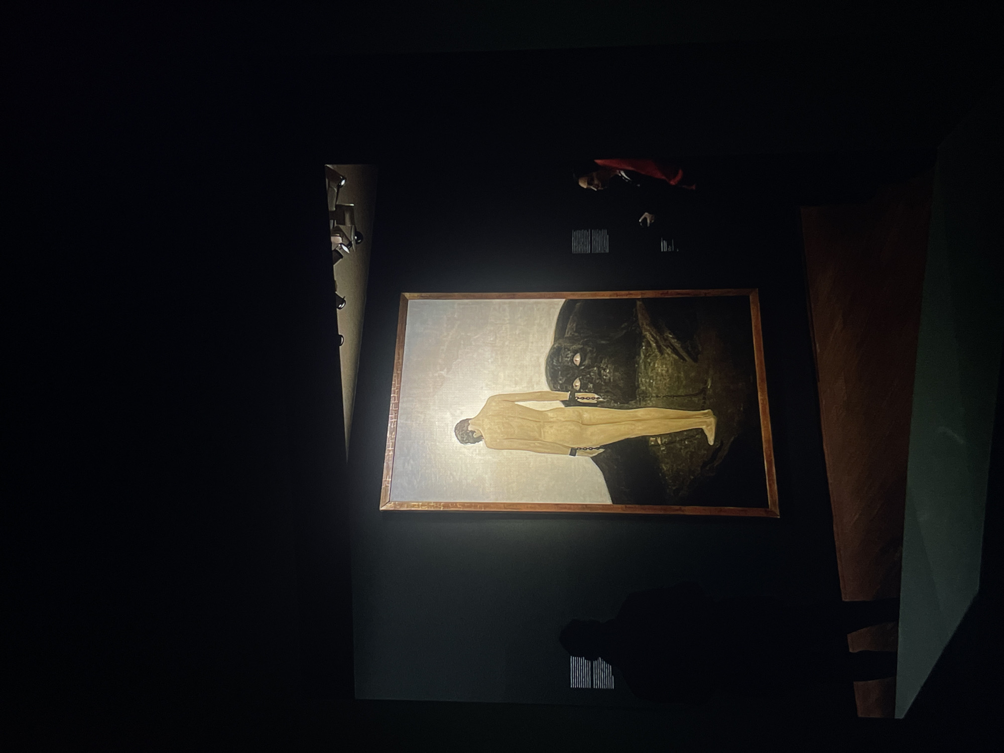 A painting of a nude male figure with what appears to be shackles on his wrists, against a dark, rocky background, hangs in a dimly lit museum. The painting is illuminated by a light source above it. The shadows of people can be seen in the room. The location is a museum in Unknown.
