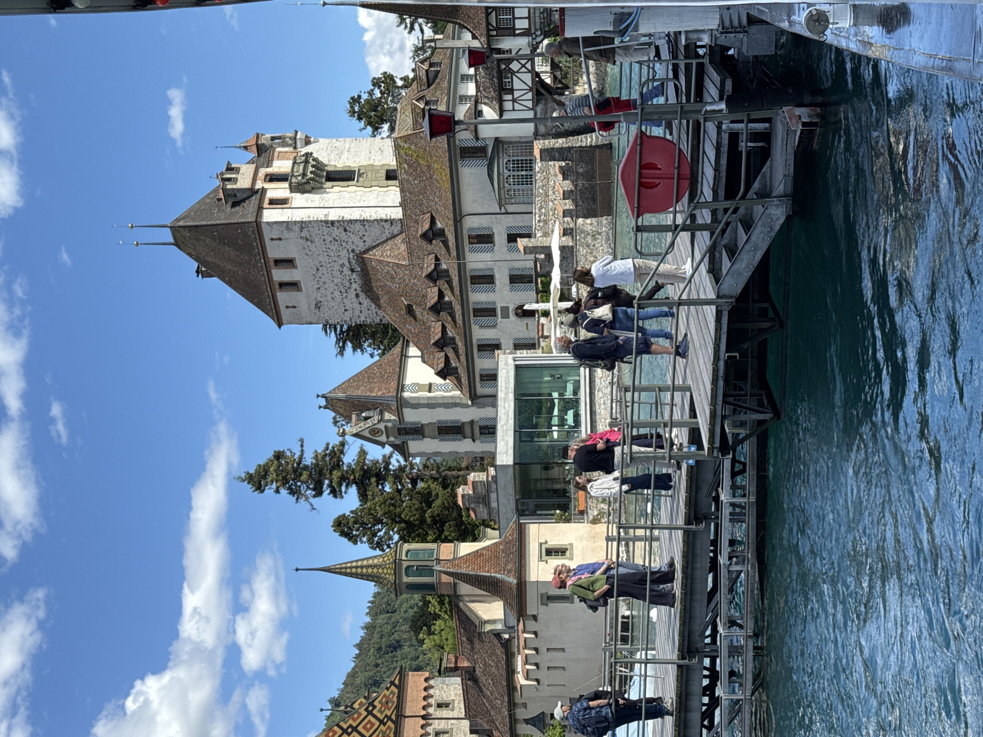 The image depicts the pier in front of Oberhofen Castle, located in Oberhofen am Thunersee, Switzerland. Several individuals are standing on the wooden pier, some appear to be tourists. The architecture of the castle is visible in the background, characterized by its distinctive towers and pitched roofs. The water in the foreground is a deep blue, suggesting a lake or river. The sky is partly cloudy with a bright blue hue. The overall scene captures a sunny day at a scenic historical site.