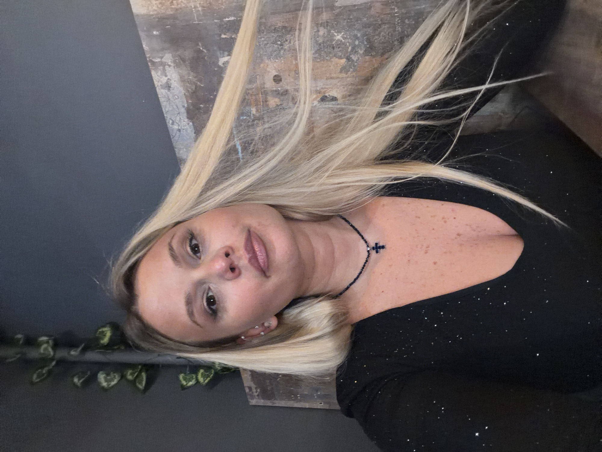 A fair-skinned woman with blonde hair is centered in the image, wearing a black, sparkling top and a dark beaded necklace with a cross pendant. Her hair is partly covering a weathered, wooden surface with stains or marks, possibly a table or wall. A gray wall with a climbing green vine is visible on the left. The image appears to be taken indoors. Sarapul, Russia is not discernible from the visual information provided.