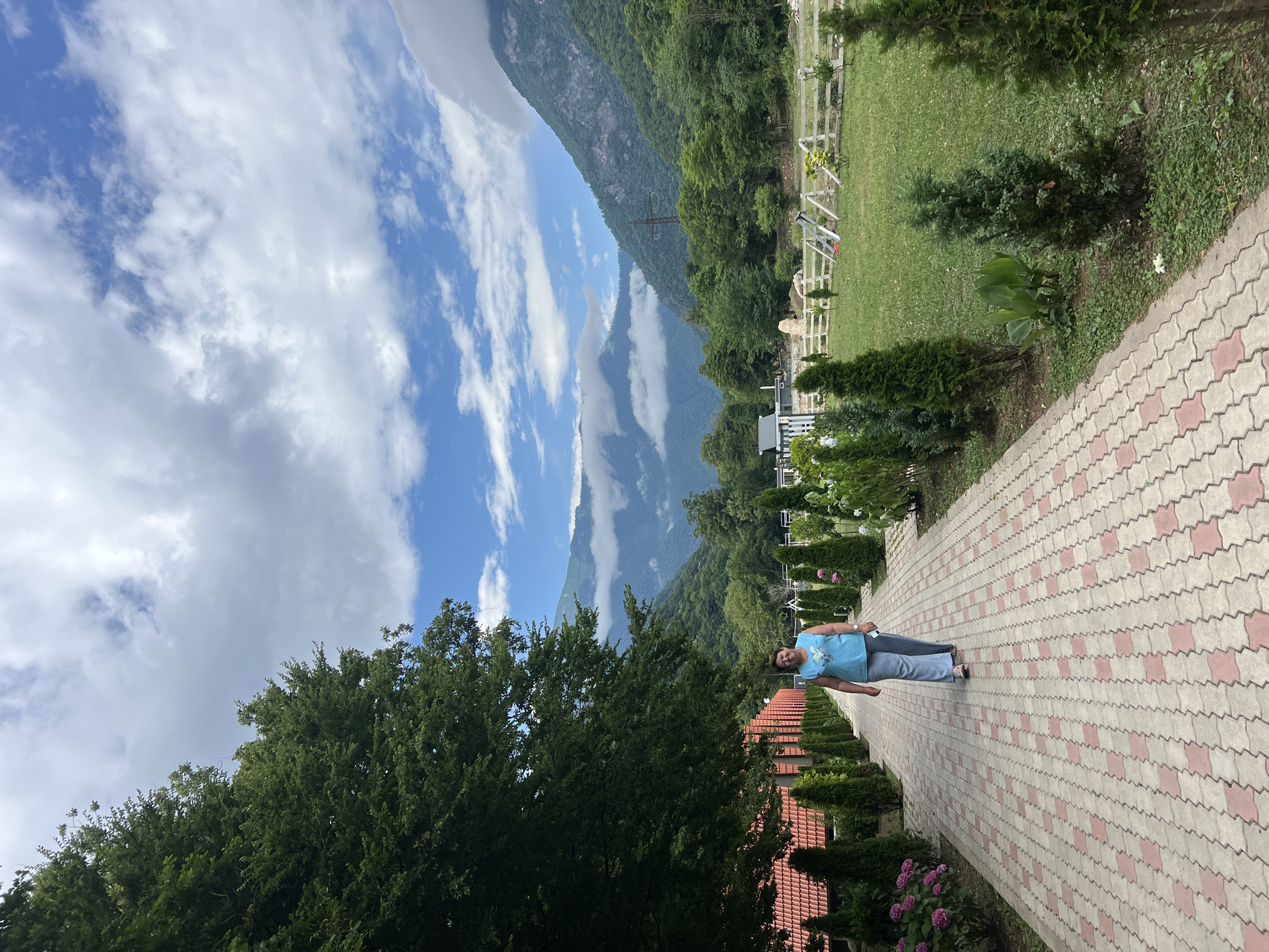 A woman is walking down a brick pathway on a sunny day. The pathway is lined with manicured trees and bushes. In the background, there are mountains and trees. The woman is wearing a blue shirt and grey pants. The sky is blue with some clouds. The overall scene is peaceful and serene.