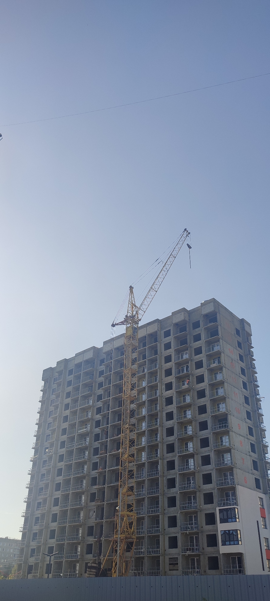 A high-rise building under construction is pictured in Novoaltaysk, Russia. A yellow construction crane is positioned next to the building, its arm extending over the top. The building itself is a concrete structure with numerous windows and balconies, many of which are open and appear unfinished. The sky above is clear and light blue. In the foreground, a grey metal fence is visible. The overall scene depicts an active construction site in a city environment.
