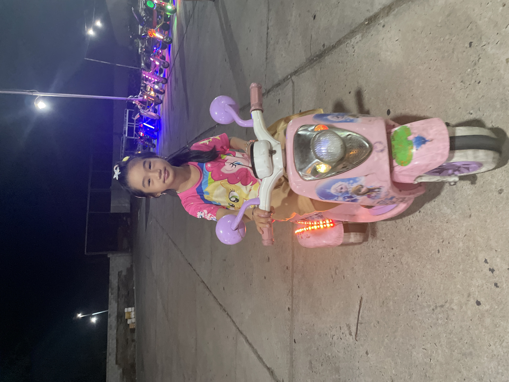 A child sits on a pink children's motorcycle in Mũi Né, Vietnam, at night. The child, wearing a pink and yellow shirt with cartoon characters, grips the handlebars of the toy motorcycle. The motorcycle has a light-up front and pink and purple details. In the background, a row of similar motorcycles with colorful lights are parked under streetlights. The ground is a concrete surface. The sky is dark. The overall scene suggests a recreational area for children's entertainment in the city.