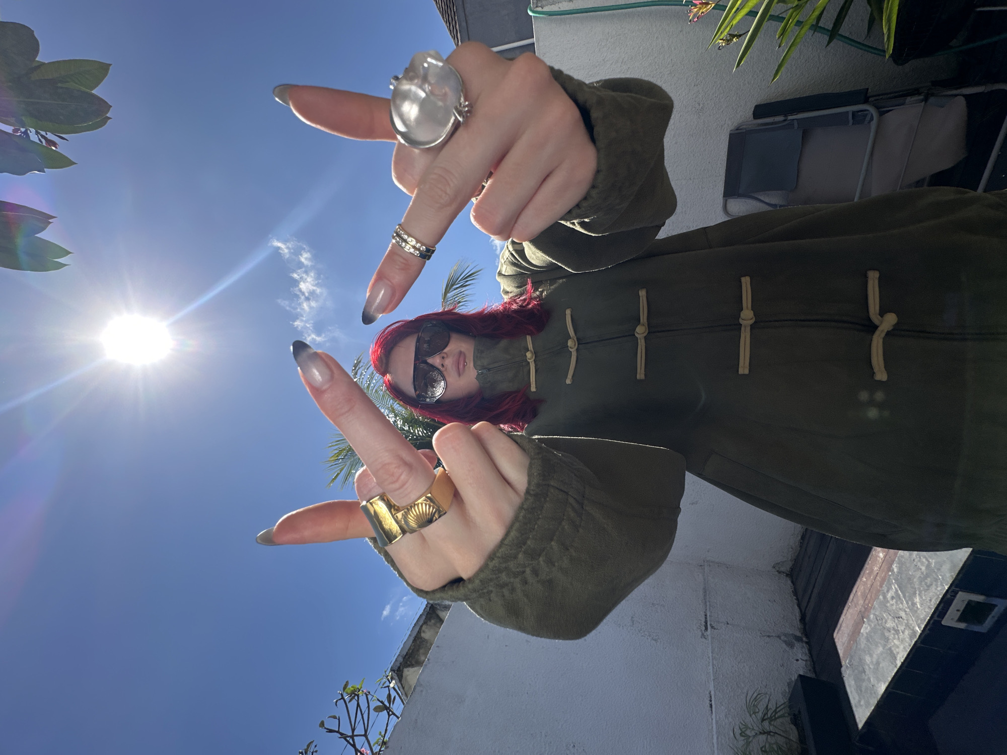 A person with bright red hair and sunglasses, wearing a green button-up coat, stands outdoors in North Kuta, Indonesia, under a bright blue sky with the sun shining. The individual is raising both middle fingers towards the camera. They are adorned with multiple rings on their fingers, including a large gold ring on the left hand and a silver ring on the right hand. The background features a white wall, some foliage, and a glimpse of a wooden deck. The person's nails are long and dark.