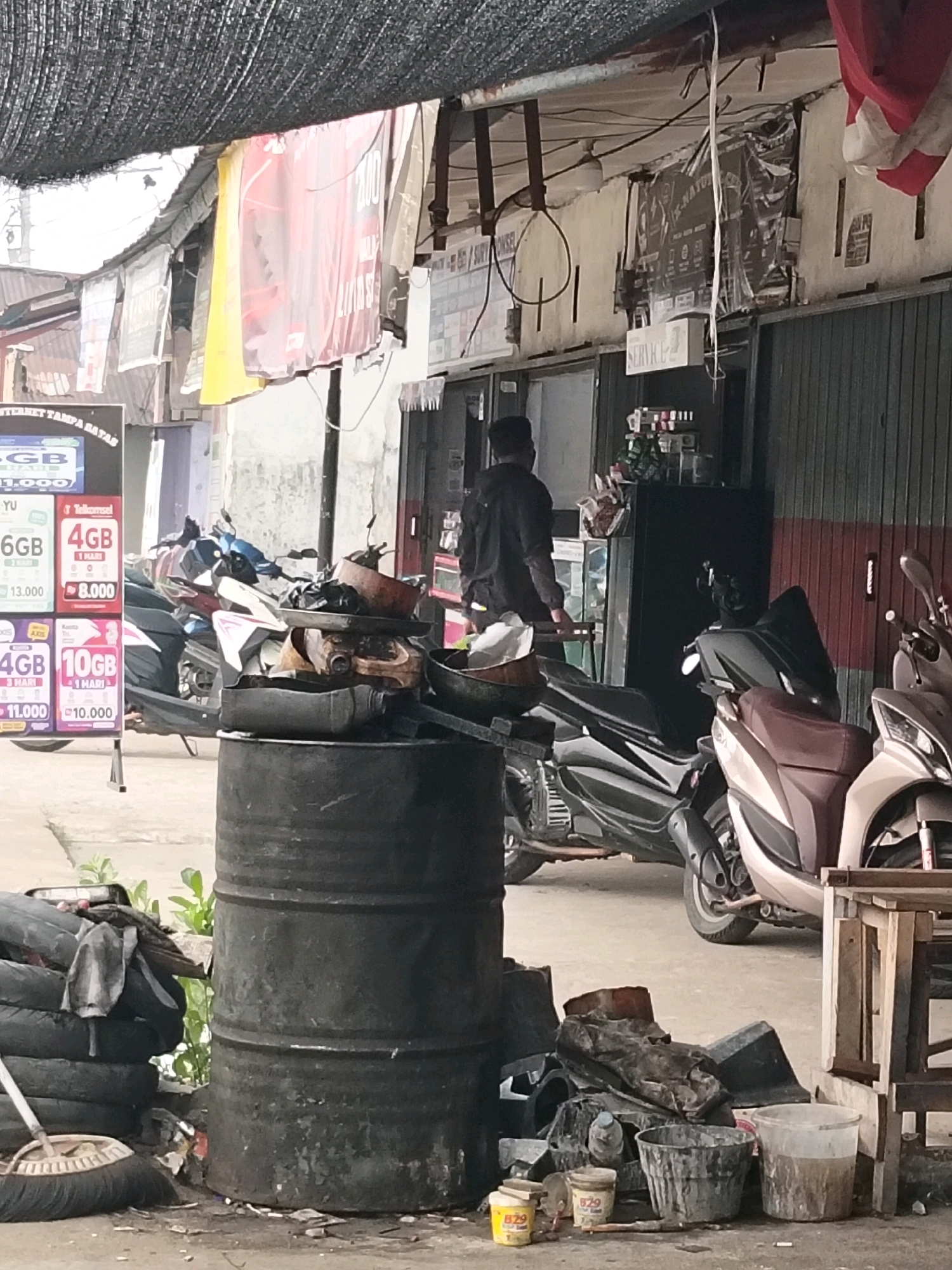In Arang Limbung, Indonesia, an outdoor scene depicts a street-level view of a commercial area. In the foreground, a large, black, weathered metal barrel sits prominently, accompanied by a pile of stacked tires and a broom. Various discarded metal objects and buckets are scattered around the barrel. Behind these items, several motorcycles are parked.
A man, dressed in a dark hooded jacket and dark pants, is visible in the mid-ground, facing away from the viewer and towards a building. He appears to be standing near a dark cabinet or refrigerator. The building behind the motorcycles displays advertising posters and banners. One poster, partially visible, features mobile data packages with prices in Indonesian Rupiah. Above the commercial space, a dark mesh structure is suspended, providing shade. The ground is paved and shows signs of dust and debris. The overall impression is one of a busy, functional, and somewhat cluttered roadside establishment, likely a motorcycle repair shop or a small retail space.