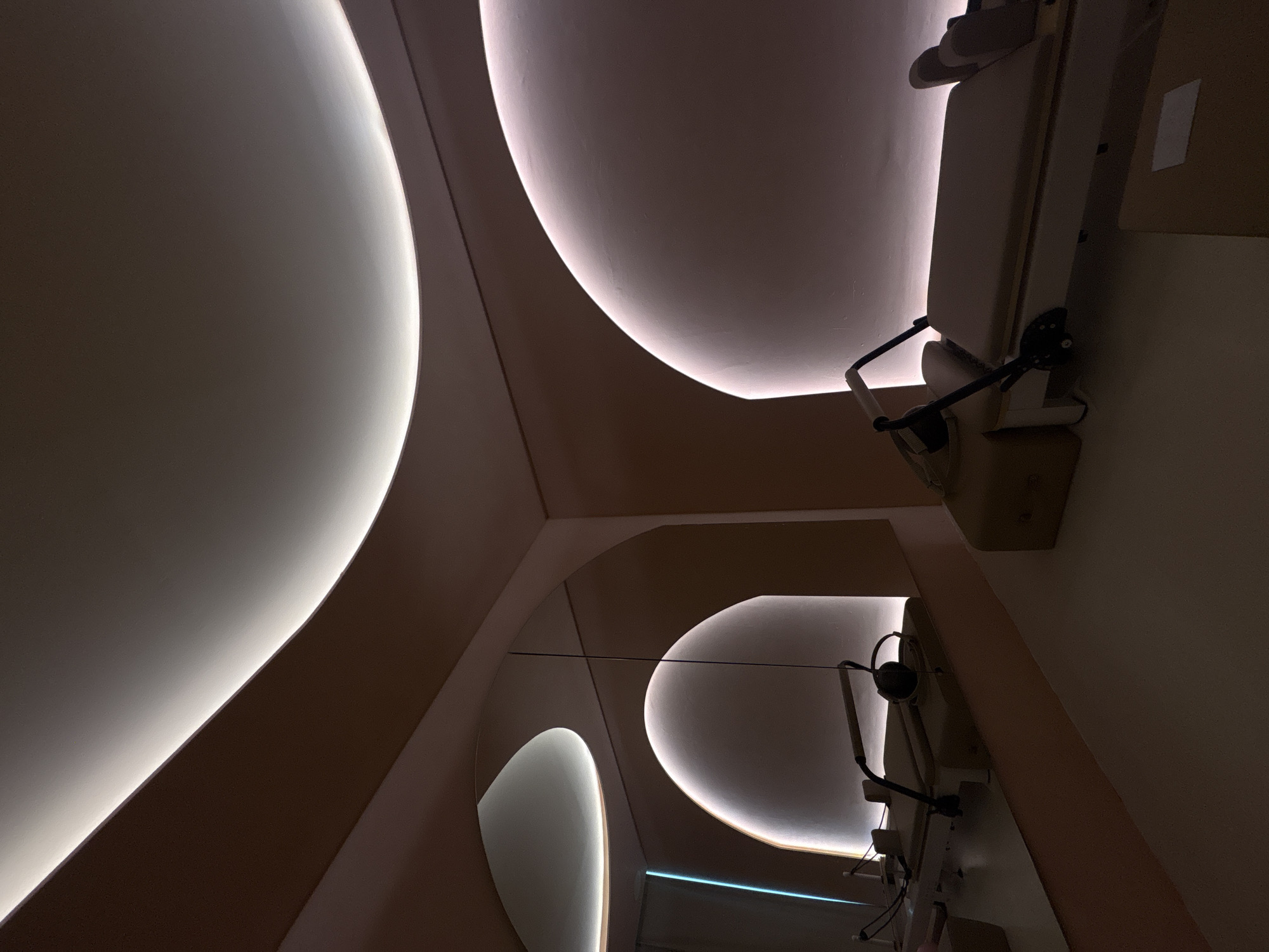 The image shows an interior space with a curved ceiling featuring integrated lighting. The walls and ceiling are light brown, and there is a partially visible object, possibly a medical examination table, mounted on the right wall. A mirror on the left reflects the curved ceiling and the object on the wall. The environment appears modern and minimalist, likely a clinic or a spa. The exact location within Mexico City cannot be determined from the image alone.