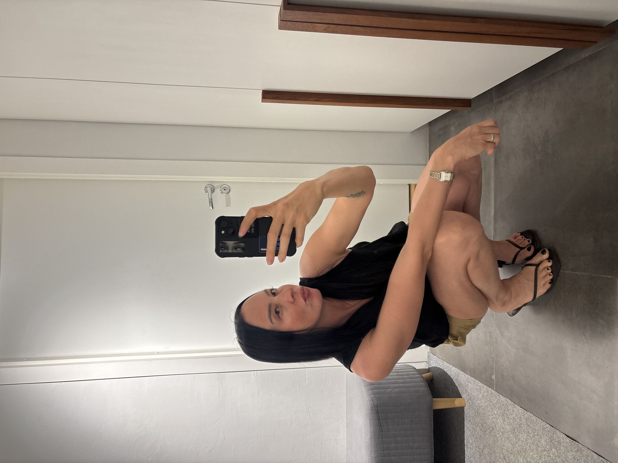 A woman is crouching and taking a selfie in what appears to be a dressing room or large closet. She is wearing a black top, tan shorts, and black flip-flops. She has dark hair and is holding a black smartphone in her right hand. She is wearing a watch on her left wrist. The background includes white wall panels with wood accents, a gray tile floor, and a gray padded bench. It is impossible to verify the exact location in the city of Chaikovsky, Russia, based solely on this image.