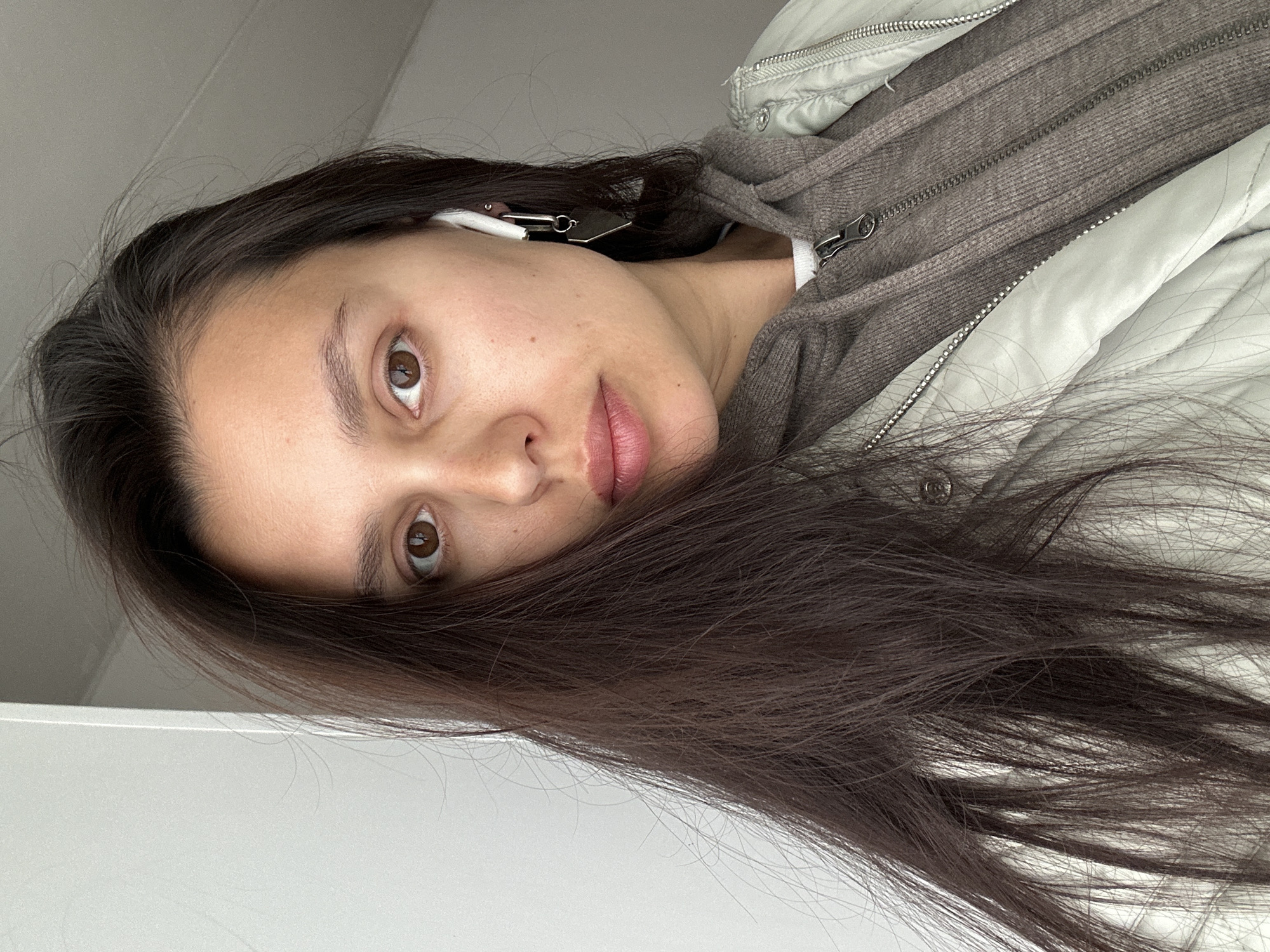 A woman with long, dark hair partially obscuring her face poses for a selfie. She is wearing a grey hoodie and a light grey jacket with zippers visible. She has fair skin, brown eyes, and pink lips. An earbud is visible in her right ear. The background is blurred and appears to be a light grey or white wall. The location is unknown and there is no information present in the image to indicate that the image was taken in Chaikovsky, Russia.