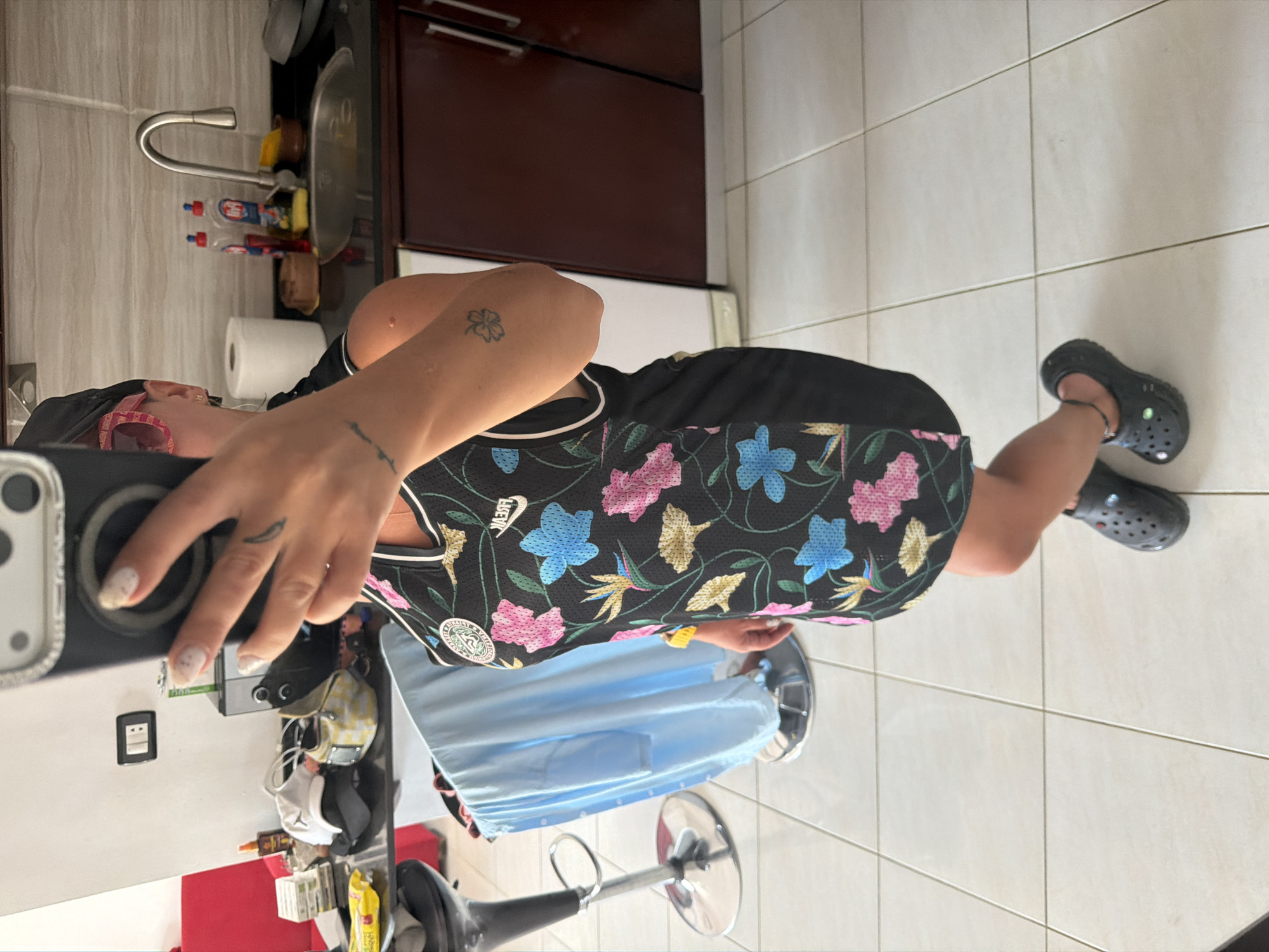 The image shows a woman standing in a room that appears to be a kitchen or dining area, possibly in Sharm El Sheikh, Egypt. She is taking a selfie with her phone. She is wearing a black dress with a floral print and dark grey Croc shoes. In the background, there is a kitchen counter with a sink and faucet, cabinets, and a roll of paper towels. There are also some objects on the counter and a red chair to the left of the frame. The floor is tiled.
