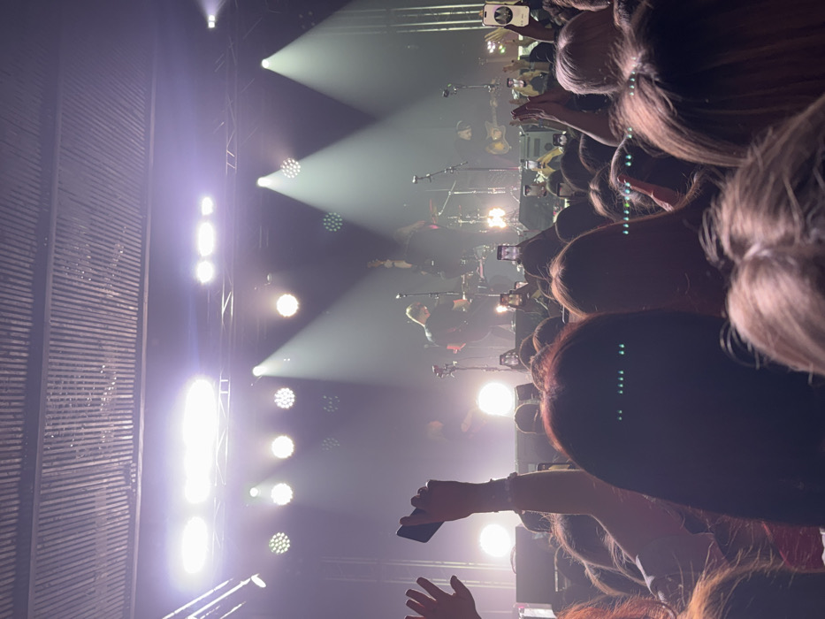 The image depicts an indoor concert scene, observed from within the audience, facing an illuminated stage. The environment includes a corrugated metal ceiling. On stage, at least four male musicians are performing, rendered partially obscured by bright stage lighting consisting of multiple spotlights and par lights casting white and purple-tinted beams through atmospheric haze. A drum kit, guitars, and microphone stands are visible musical equipment. The audience, primarily in the foreground and midground, consists of numerous individuals with various hair colors and styles, many of whom are holding up smartphones to record the performance, and several hands are raised. This scene is located in Izhevsk, Russia.