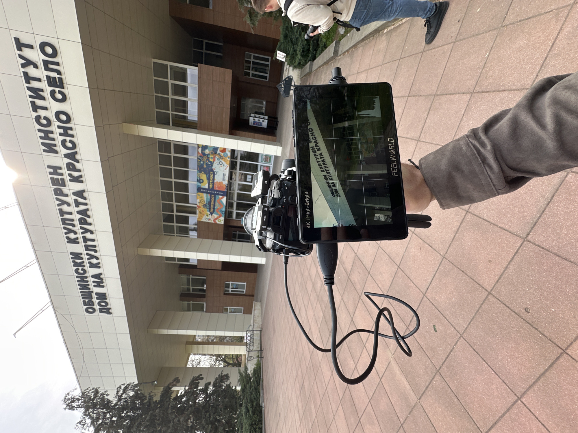 The image shows a camera with a monitor attached, pointed towards the front of a building. The building is identified by signage in Bulgarian as "OBSHTINSKI KULTUREN INSTITUT DOM NA KULTURATA KRASNO SELO," which translates to "MUNICIPAL CULTURAL INSTITUTE HOUSE OF CULTURE KRASNO SELO." This indicates the location is likely in Krasno Selo, a district in Sofia, Bulgaria. The monitor displays the image being captured by the camera, showing the front of the building. One person is seen holding the camera, while another person is partially visible in the background. The ground appears to be paved.