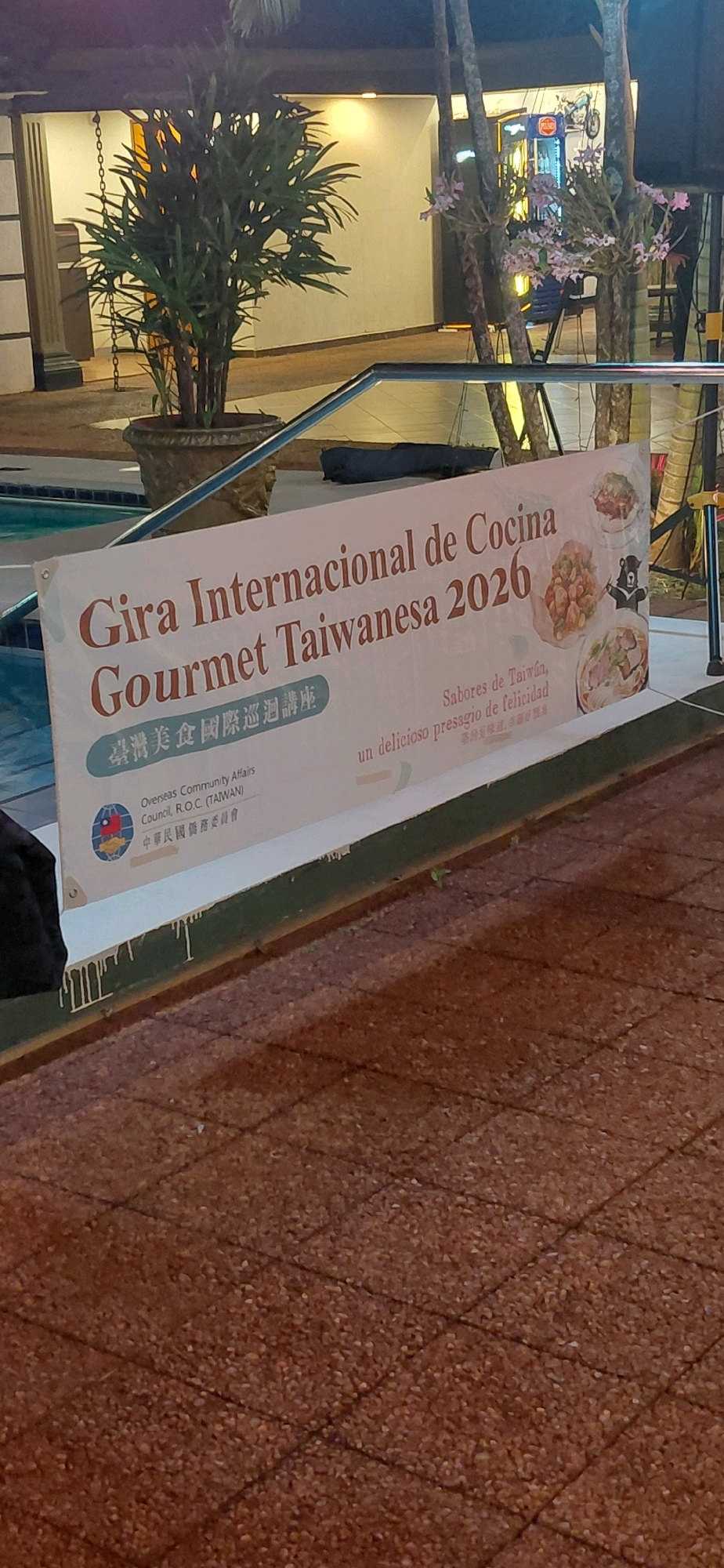 The image depicts a banner advertising a "Gira Internacional de Cocina Gourmet Taiwanesa 2026" (International Gourmet Taiwanese Cuisine Tour 2026). The banner is predominantly white with text in red and brown. It features images of Taiwanese food and a depiction of a Taiwanese black bear. At the bottom left, there is a logo and text that reads "Overseas Community Affairs Council, R.O.C. (TAIWAN)" and Chinese characters. To the right, there is text in Spanish, "Sabores de Taiwán, un delicioso presagio de felicidad."
The banner appears to be displayed outdoors, possibly near a body of water such as a swimming pool, suggested by the blue edge visible on the left. Behind the banner, there are tropical plants in pots and trees with some flowering branches. In the background, a building with outdoor lighting is visible. The foreground shows a textured ground surface, likely a paved area. The overall lighting suggests it might be evening or nighttime.
The image was taken in Encarnación, Paraguay, likely at a venue hosting this culinary event. The presence of the "Overseas Community Affairs Council, R.O.C. (TAIWAN)" indicates a cultural exchange or promotional event organized by Taiwan.