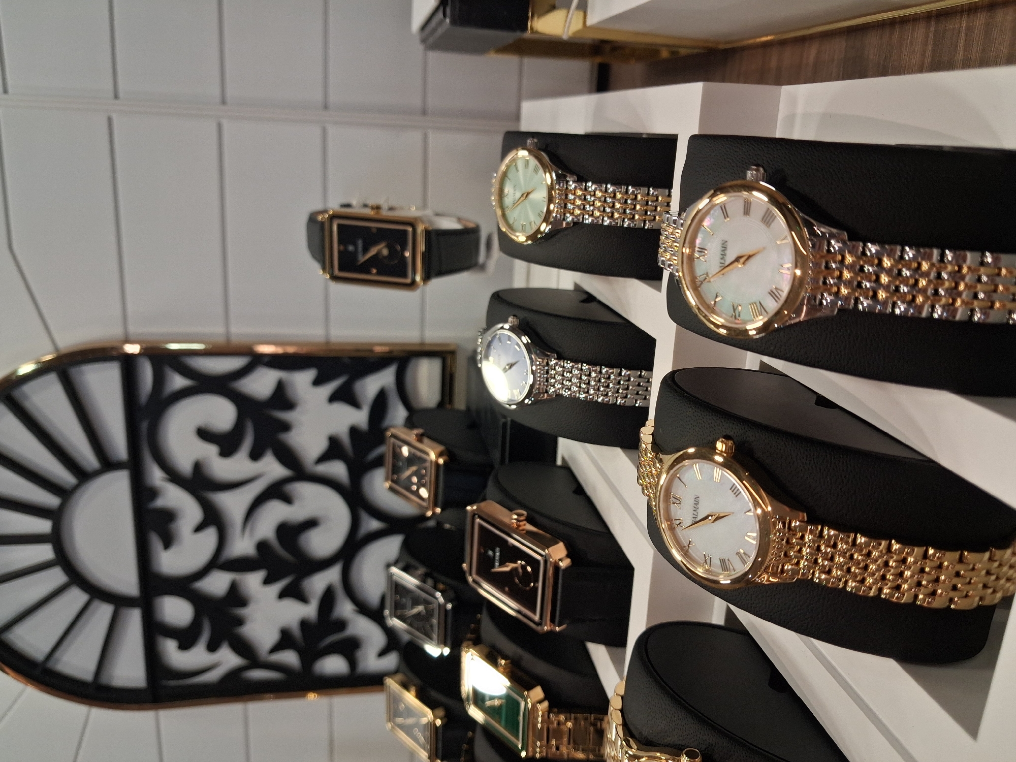 The image depicts a display of Balmain watches in what appears to be a retail store or showroom. The watches, a mix of rectangular and round face designs with varying bands (leather, silver, and gold), are presented on black, cylindrical display stands. A decorative metal grille with a sunburst and scroll pattern is visible to the left. The lighting suggests an indoor environment, possibly a jewelry or department store. While specific location in Varna, Bulgaria, isn't discernible, the style and content suggest a shop catering to luxury goods.