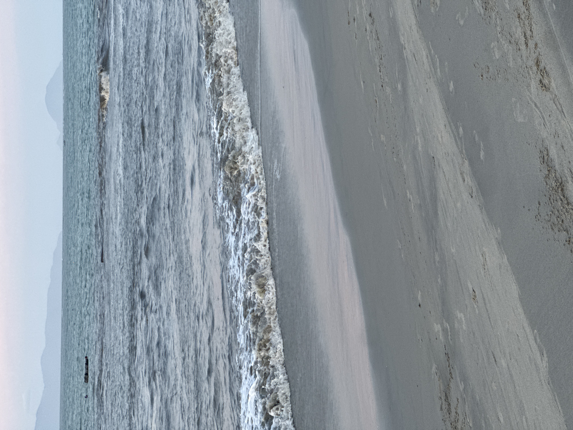 The image shows a beach in Đà Nẵng, Vietnam. The camera is oriented looking down the beach. The sea is on the left side of the image, with waves crashing onto the sand. The sand is a light gray color, and there are some darker patches of sand. A portion of mountains are seen in the very far left background. In the sea in the middle-left of the frame are a few objects that appear to be standing upright. The sky is overcast.