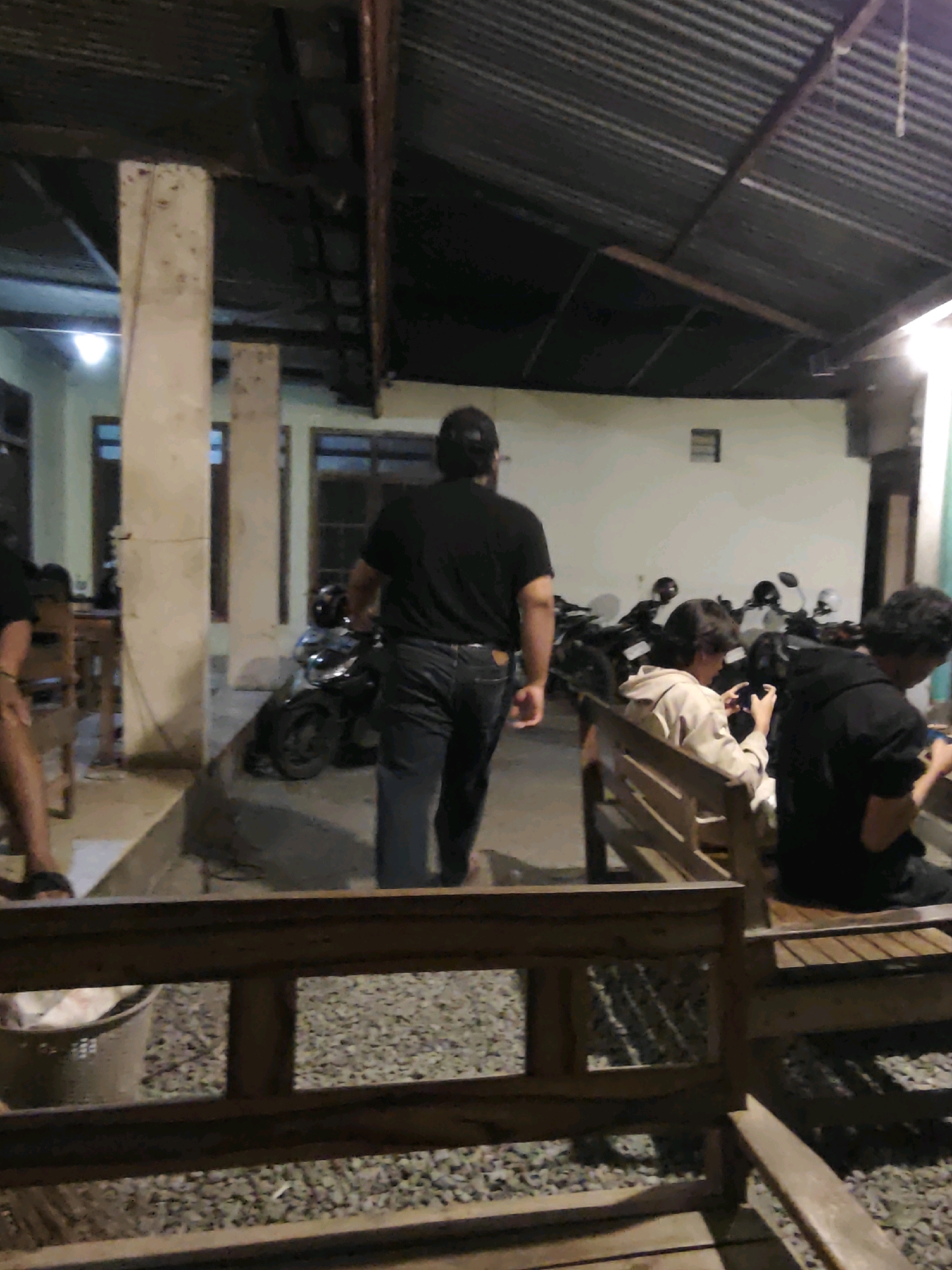 In Ngasem, Indonesia, an outdoor gathering area features several people. In the foreground, a wooden bench is visible, with gravel on the ground. To the left, a person's legs and feet are seen, wearing sandals. In the mid-ground, a man wearing a black t-shirt and jeans walks away from the camera. Behind him, a motorcycle is parked. Further back, several other motorcycles are lined up. Two individuals are seated on a wooden bench in the mid-ground, both looking at their phones. One person wears a light-colored hooded jacket, and the other wears a black hooded jacket. The environment appears to be an open-air shelter with a corrugated metal roof supported by concrete pillars. The wall behind the motorcycles is white, and windows are visible. The lighting suggests it is evening or nighttime.