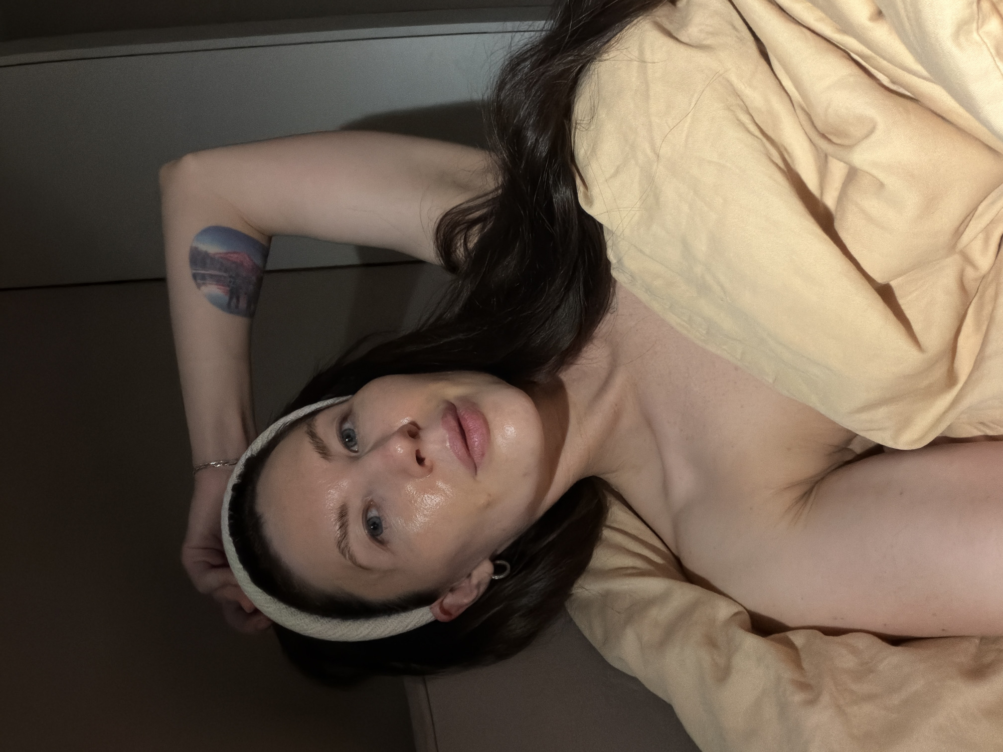 A woman is lying down, partially covered by a tan-colored blanket or sheet. She is wearing a cream-colored headband. Her dark hair is loose. She has a tattoo on her upper arm. The location is likely indoors. There is nothing in the image to indicate the city of Perm, Russia.