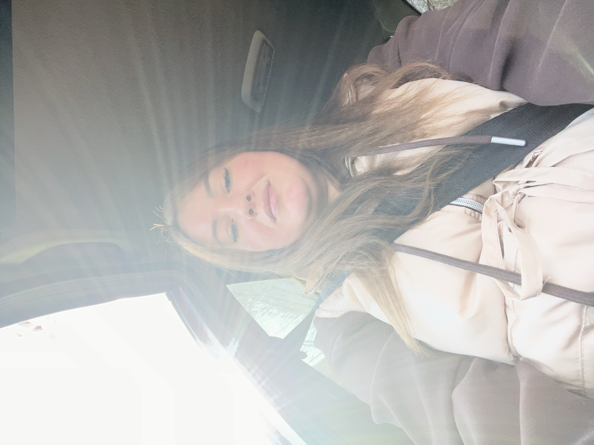 The image depicts a woman with light brown hair wearing a beige jacket and a dark seatbelt. She is sitting in a car, possibly a passenger, with a bright light source visible through the window, obscuring the view. The location is suggested as Chaikovsky, Russia, based on information provided, but there are no visual clues in the image to confirm the specific location. The woman is facing the camera and smiling slightly. The car's interior and the woman's clothing are the only discernible objects. No specific action or interaction is evident.