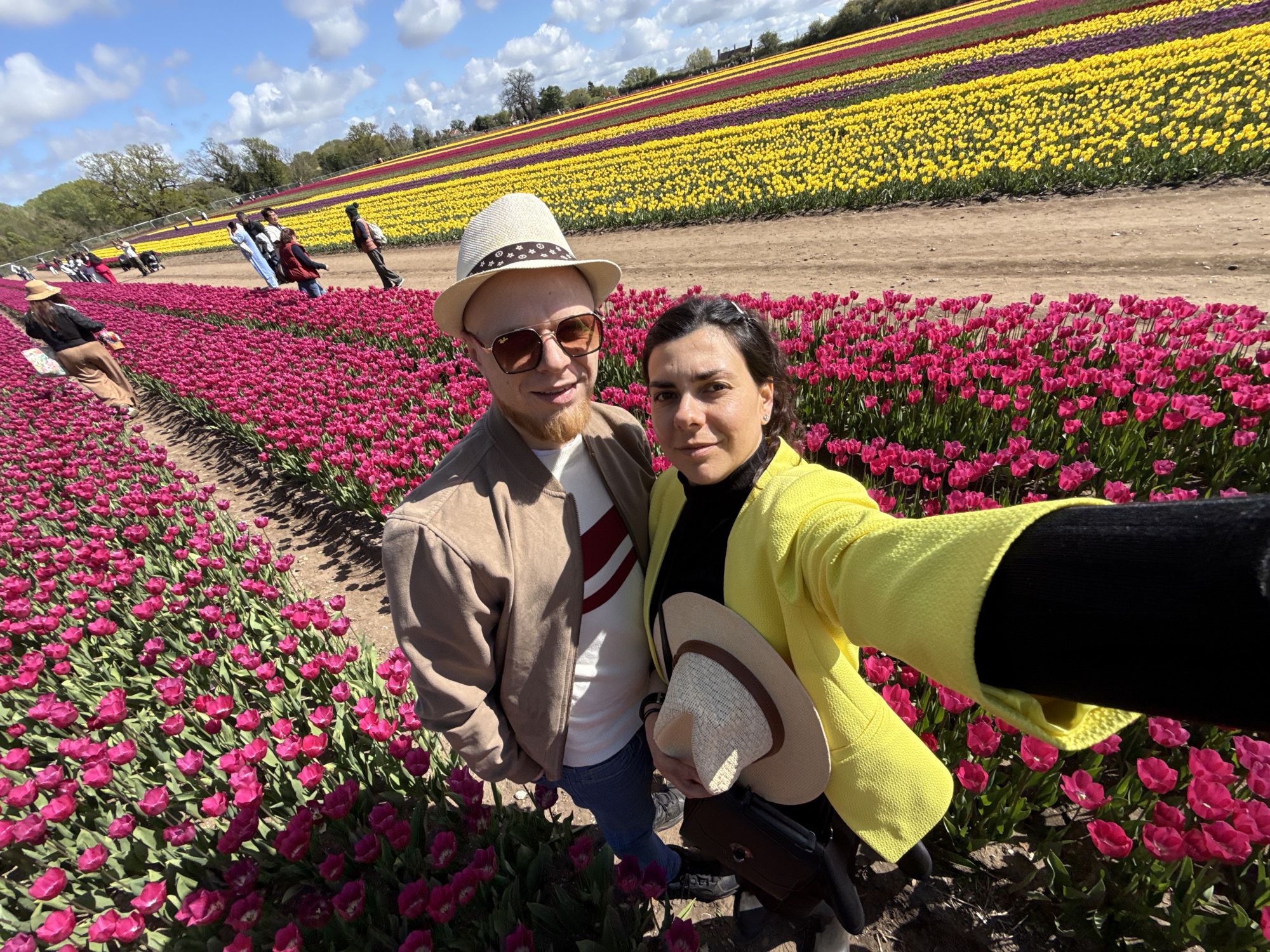 A man and a woman are captured in a selfie, posing in a vibrant tulip field under a bright, partly cloudy sky. The man, on the left, wears a beige jacket over a white top with red stripes, blue jeans, and dark sunglasses, complemented by a light-colored fedora-style hat. He has a beard and a slight smile. The woman, on the right, sports a bright yellow textured blazer over a black top, and holds a natural-colored fedora hat in her hand. Her dark hair is styled with small clips, and she offers a pleasant expression to the camera.
They are situated amidst numerous rows of vivid pink tulips that fill the immediate foreground and extend towards the mid-ground. Beyond these, the extensive field stretches into the distance, displaying distinct, parallel stripes of different colored tulips, including bright yellow, purple, and deeper red varieties, creating a striking panoramic pattern. Dirt paths separate the various tulip rows. Several other people are visible in the background, walking leisurely along these paths, enjoying the scenic floral display. Trees line the distant horizon beneath a clear blue sky dotted with fluffy white clouds, suggesting a sunny and pleasant day. The image captures a lively scene at a tulip farm or festival, likely in King's Lynn and West Norfolk, United Kingdom. No text is visible in the image.