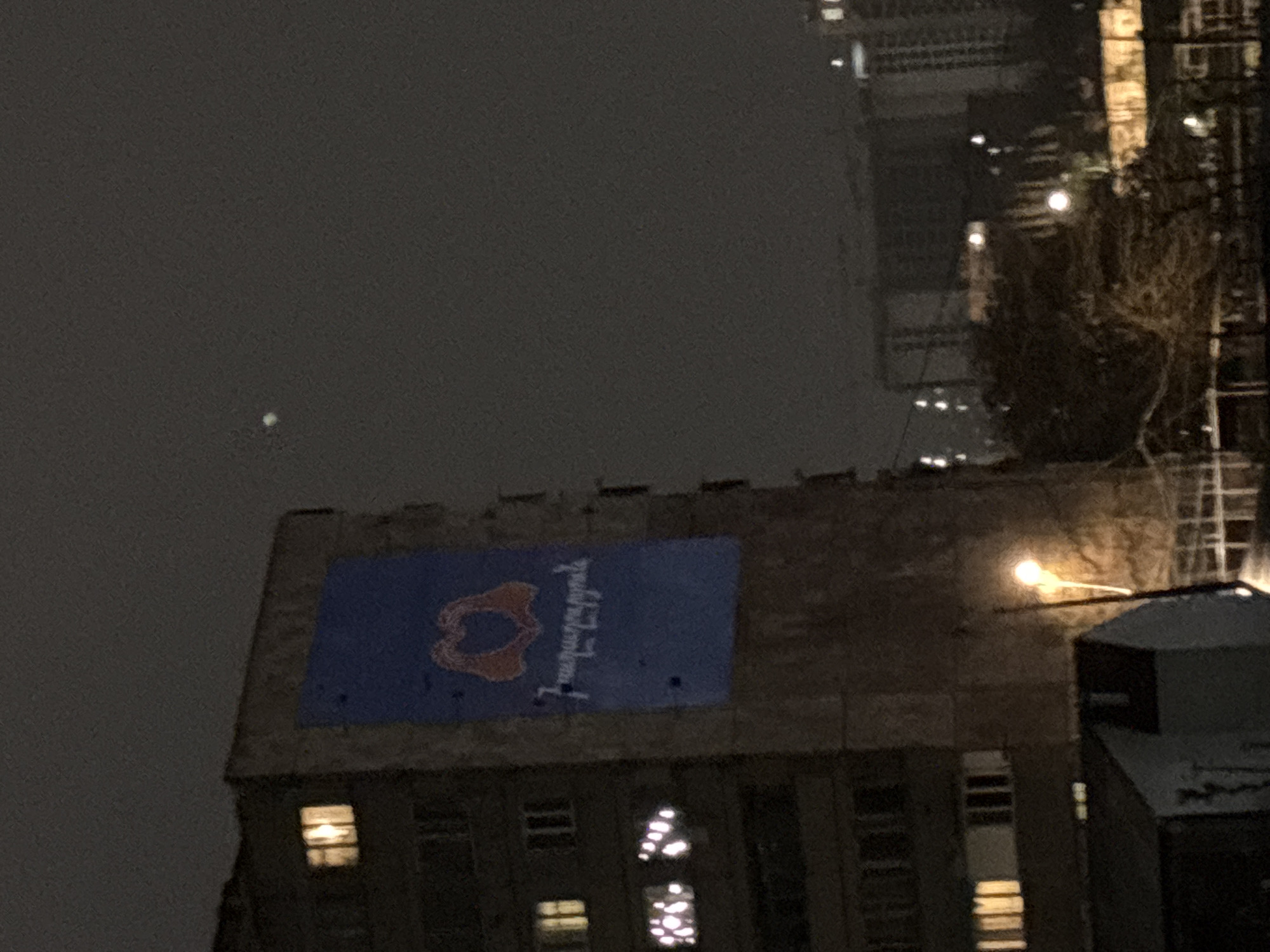 The image shows a building in Yerevan, Armenia at night. The building has a large blue banner on its roof with an image of the Artsakh flag and Armenian script. The text reads, "Artsakh is Armenia. Period." The sky is dark and cloudy. Other buildings are visible in the background, along with streetlights.