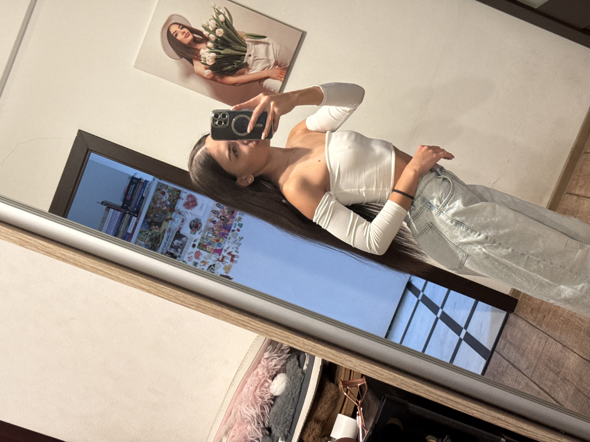 The image depicts a young woman in a white off-the-shoulder crop top and light-wash wide-leg jeans, taking a mirror selfie. She is holding a smartphone in her right hand, with her left hand resting on her hip. Her long dark hair cascades down her back. In the background, a large mirror reflects the room, showing a checkered floor, a wooden floor, and various decorative items on shelves, including books and framed pictures. A framed picture of a woman holding tulips is visible on the wall. The scene appears to be an indoor setting, possibly a bedroom or dressing area. There is no indication of the specific location within Chișinău, Moldova.
