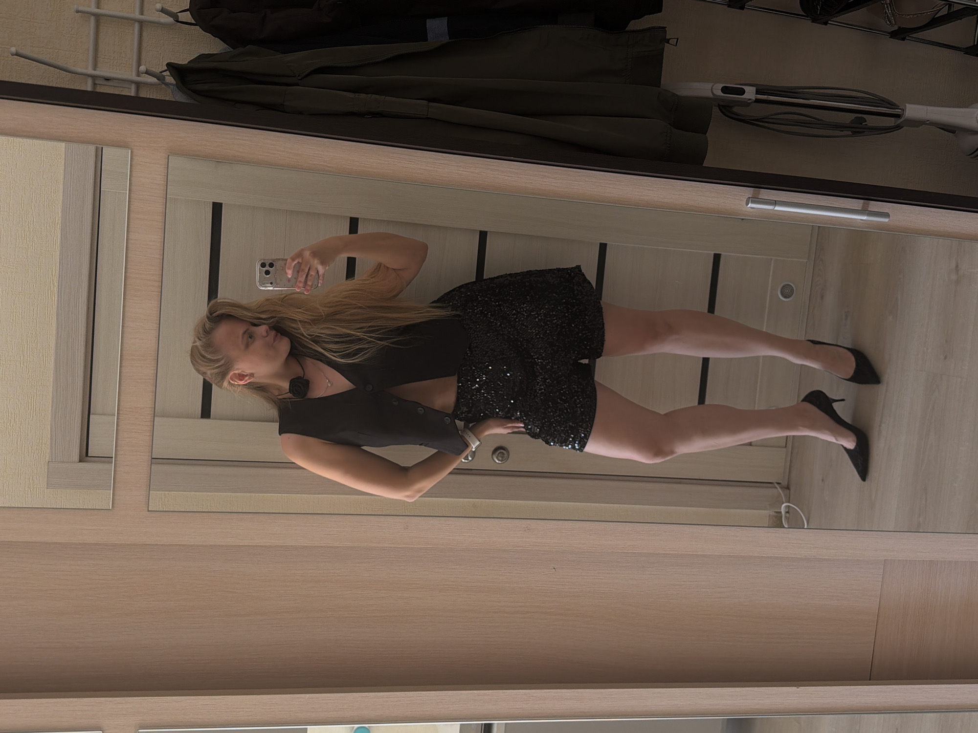 A woman is taking a mirror selfie in what appears to be a room with light wood-paneled walls and a mirrored closet. She is wearing a black dress, black vest, and black high heels. She is holding a smartphone. The background shows a closet with clothes hanging inside and a vacuum cleaner stored on the top shelf. There is no clear indication of the city of Izhevsk, Russia.