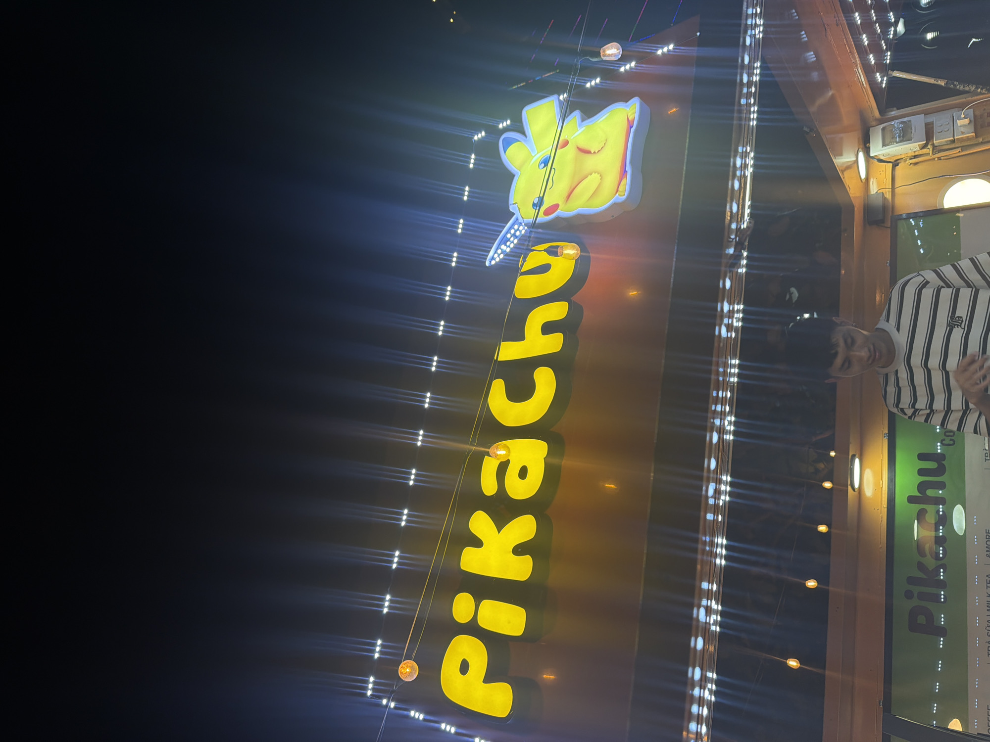 The image depicts a nighttime scene in Nha Trang Ward, Vietnam. A bright, vertical sign displays "Pikachu" in yellow, with an animated Pikachu figure at the top. The sign is illuminated by decorative lights, and a person is partially visible on the right side of the frame.