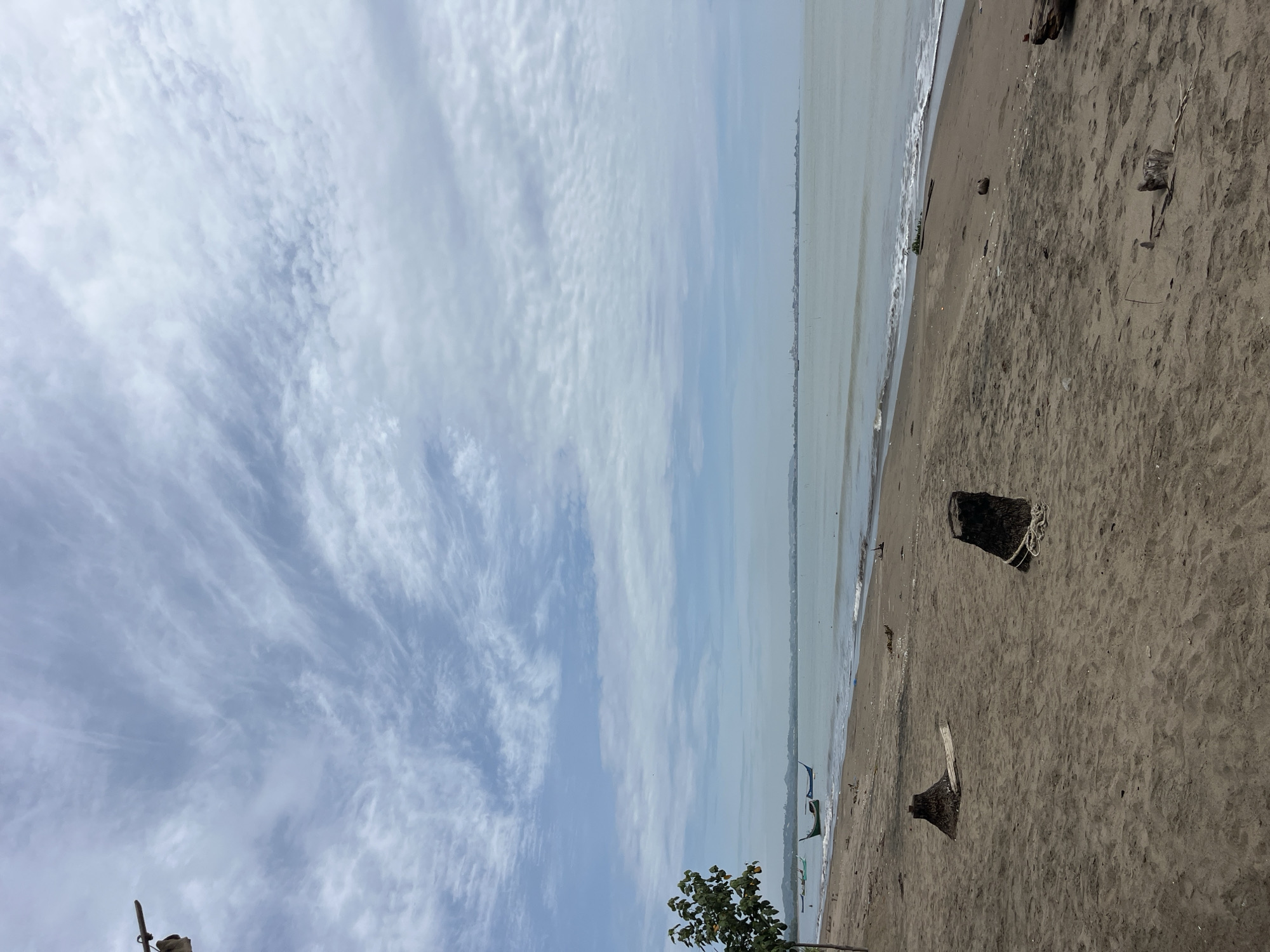 The image shows a beach scene in Lhokseumawe, Indonesia. The sky is predominantly blue with wispy, white clouds. The sea is a murky grey-green, with gentle waves lapping at the sandy shore. The sand is a dark grey color and scattered with debris, including tree stumps and driftwood. In the distance, a faint skyline of buildings is visible. In the foreground, there is a small tree. Several fishing boats are visible on the horizon.