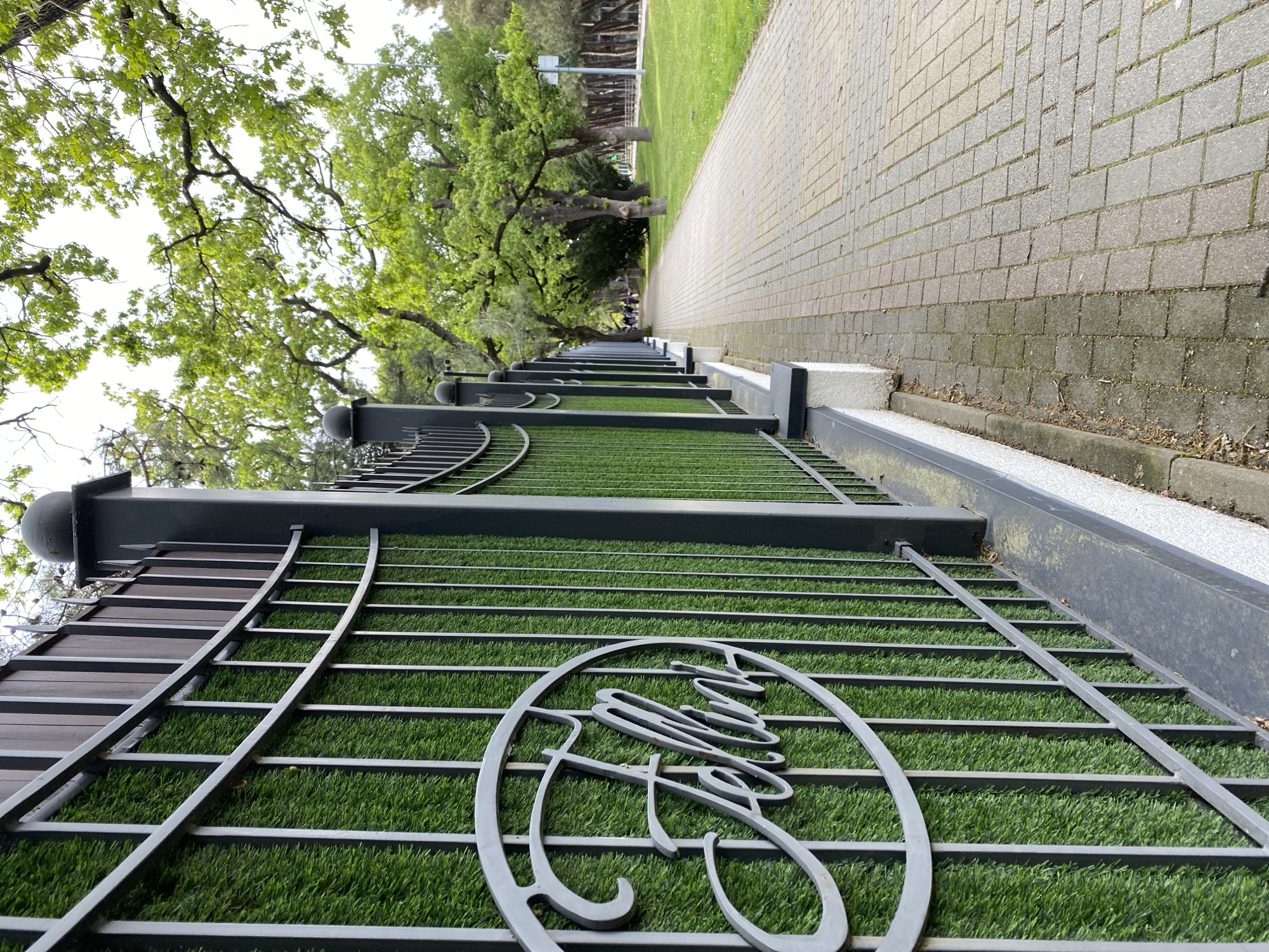 This image depicts a public park or promenade in Gelendzhik, Russia, viewed as if rotated 90 degrees clockwise from its original orientation.
In the foreground, a dark grey metal fence or barrier runs along the right side of the frame. This fence features horizontal bars and decorative, curving elements. Behind the fence, visible through its structure, is a strip of vibrant green artificial turf. A prominent detail on the lower part of the fence is a stylized, metallic logo or word in a cursive script, which appears to be "Gelendzhik." The fence is anchored on a light-colored concrete base.
To the left of the fence, a paved pathway made of light grey rectangular bricks stretches into the distance, suggesting a long promenade. Adjacent to the path, on its left, is an area of natural green grass. Further left, mature trees with abundant green foliage line the pathway, forming a lush canopy overhead.
In the far distance along the pathway, several small human figures are discernible, indicating people are using the walkway. The sky, visible through the tree branches, is bright and overcast, suggesting it is daytime on a cloudy day, with soft, diffused lighting. No specific activities are clearly happening beyond people walking in the distance.