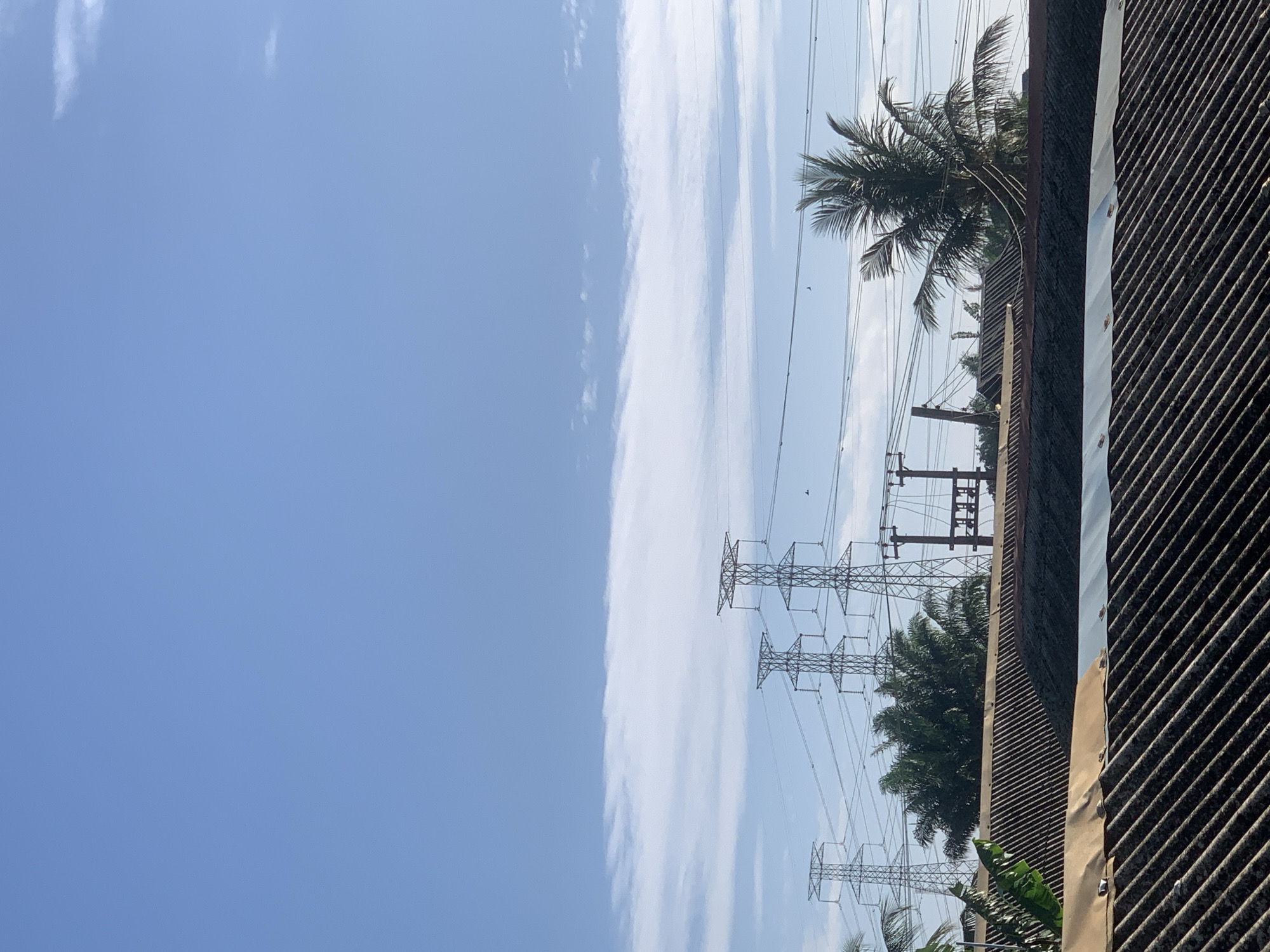 The image shows a view in Ikorodu, Nigeria, featuring a blue sky with wispy white clouds dominating the left side of the frame. Power lines stretch across the scene, supported by metal pylons. Palm trees are interspersed among the pylons and lines. To the right of the power lines, a corrugated metal roof is visible, providing a ground-level perspective. The scene appears to be from a lower vantage point looking up, capturing a mix of urban infrastructure and natural elements.
