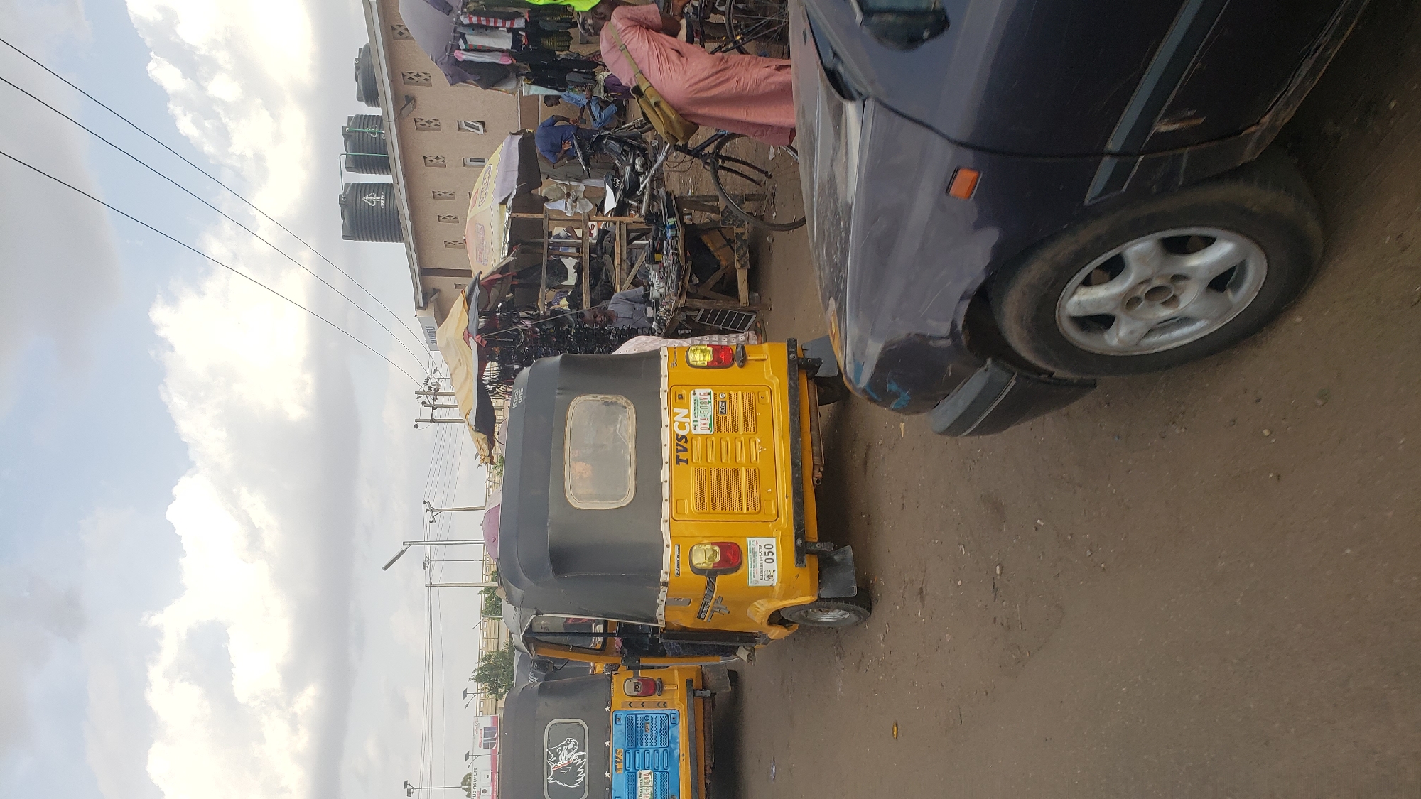 The image depicts a street scene in Kawo, Nigeria. A yellow-and-black tricycle, commonly known as a "Keke Napep", is parked in the street. To the right, the front quarter of a dark blue car is visible. In the background, there are shops and stalls selling various items, with people milling about, including one person on a bicycle and dressed in light pink robes. The sky is overcast with some visible clouds, and the road surface appears to be paved. Three black water tanks can be seen on top of a building. Overhead power lines run across the top of the frame.