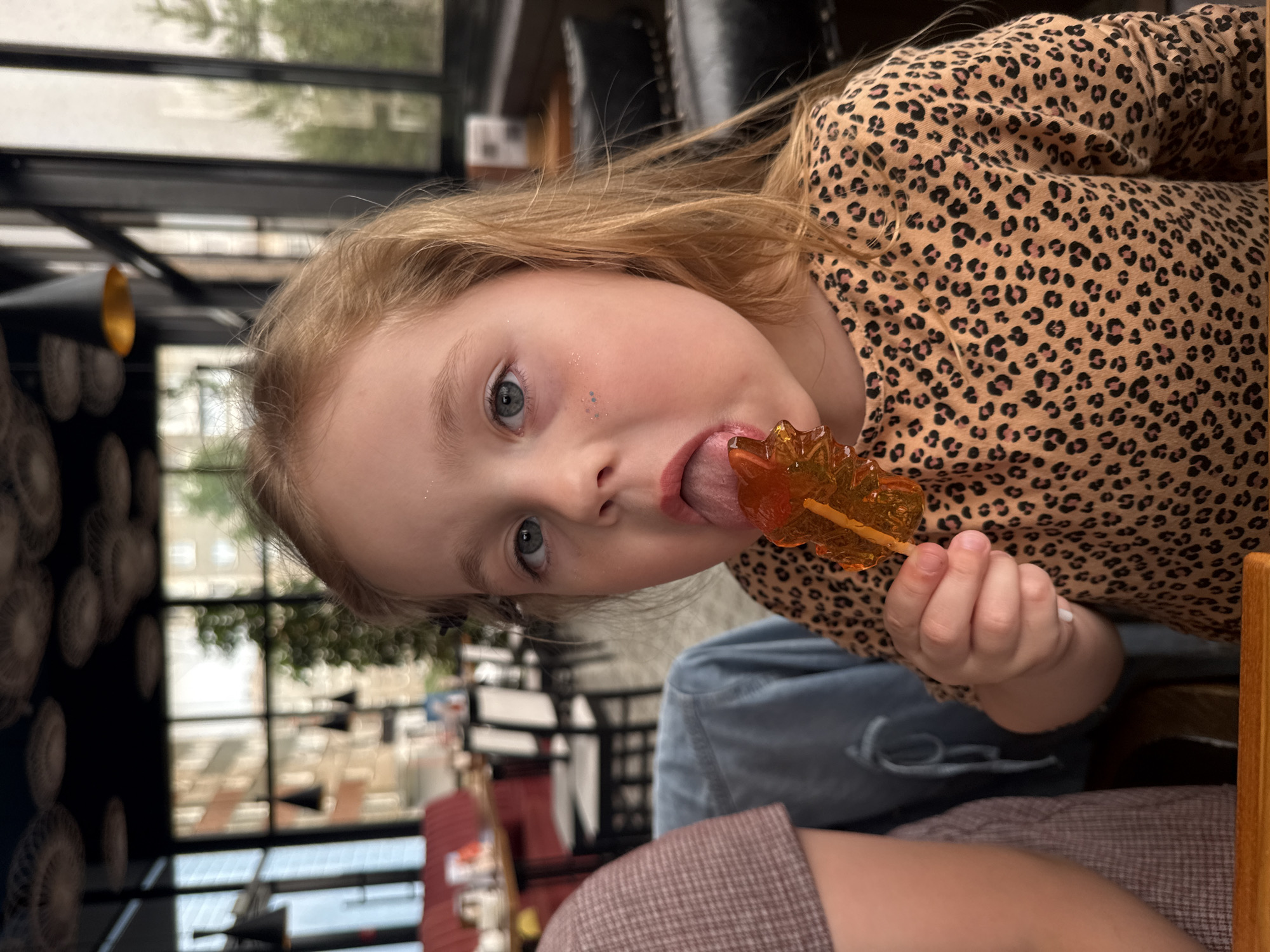 A young girl with blonde hair is pictured in a close-up shot, holding and licking a translucent orange lollipop shaped like a leaf. She has light blue eyes and is wearing a long-sleeved shirt with a leopard print pattern. She has some glitter on her cheek. The background suggests an indoor setting, possibly a cafe or restaurant, with large windows revealing an urban exterior. The image focuses on the girl and her treat, with the background blurred, hinting at a city environment, but the exact location within Izhevsk, Russia is not discernible from the visual information alone.