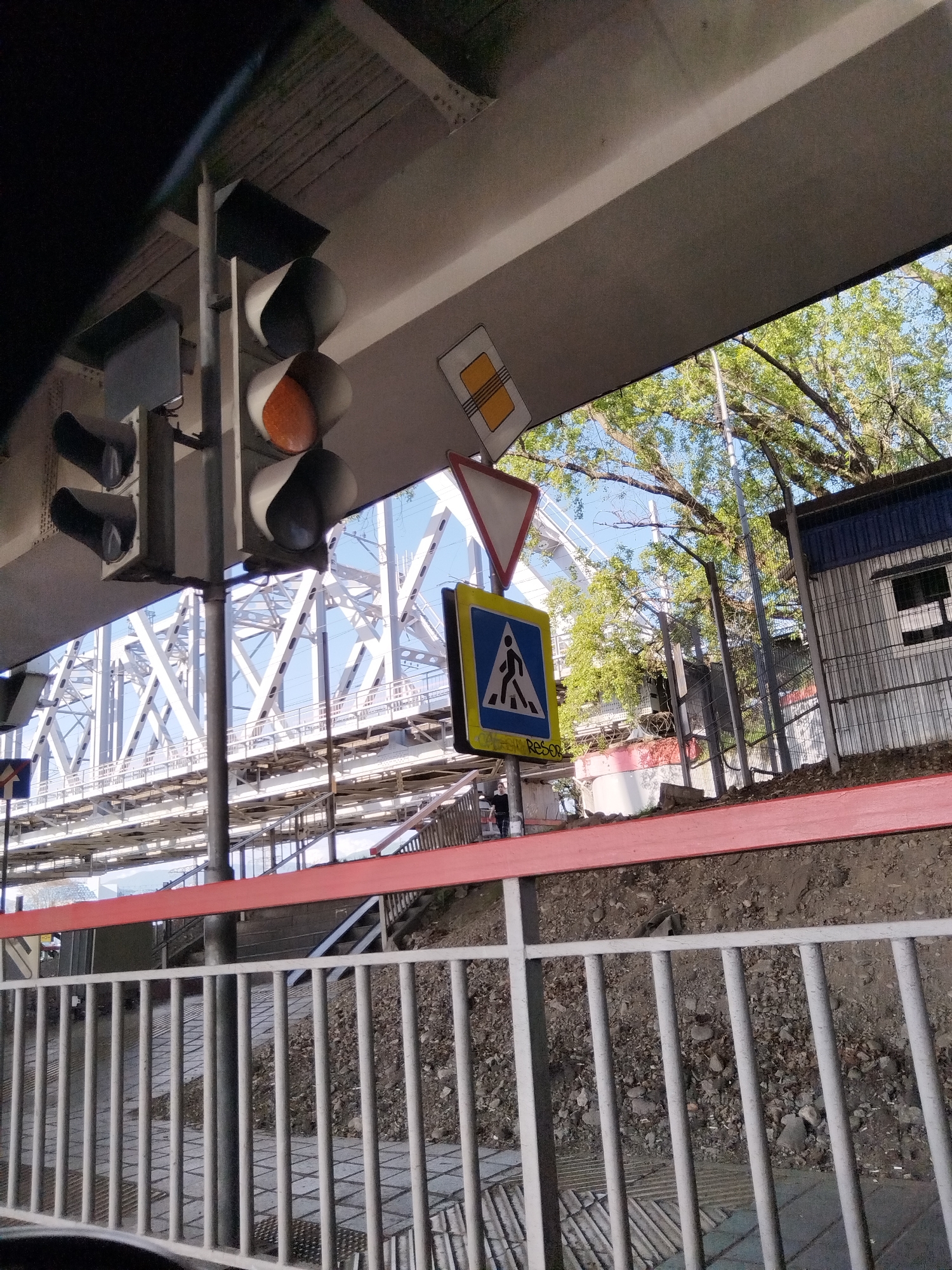 The image depicts a scene in Sochi, Russia, near a railway bridge. The view is framed by an overpass structure, revealing elements of the bridge's white steel framework. In the foreground, there is a set of traffic signals with an orange light illuminated. Several road signs are visible, including a pedestrian crossing sign. A metal guardrail separates the lower ground from a raised embankment, which is marked with a red band. The background includes a small building or shed with a blue roof, trees with green foliage, and sections of a security fence. The image appears to be taken from inside a vehicle.