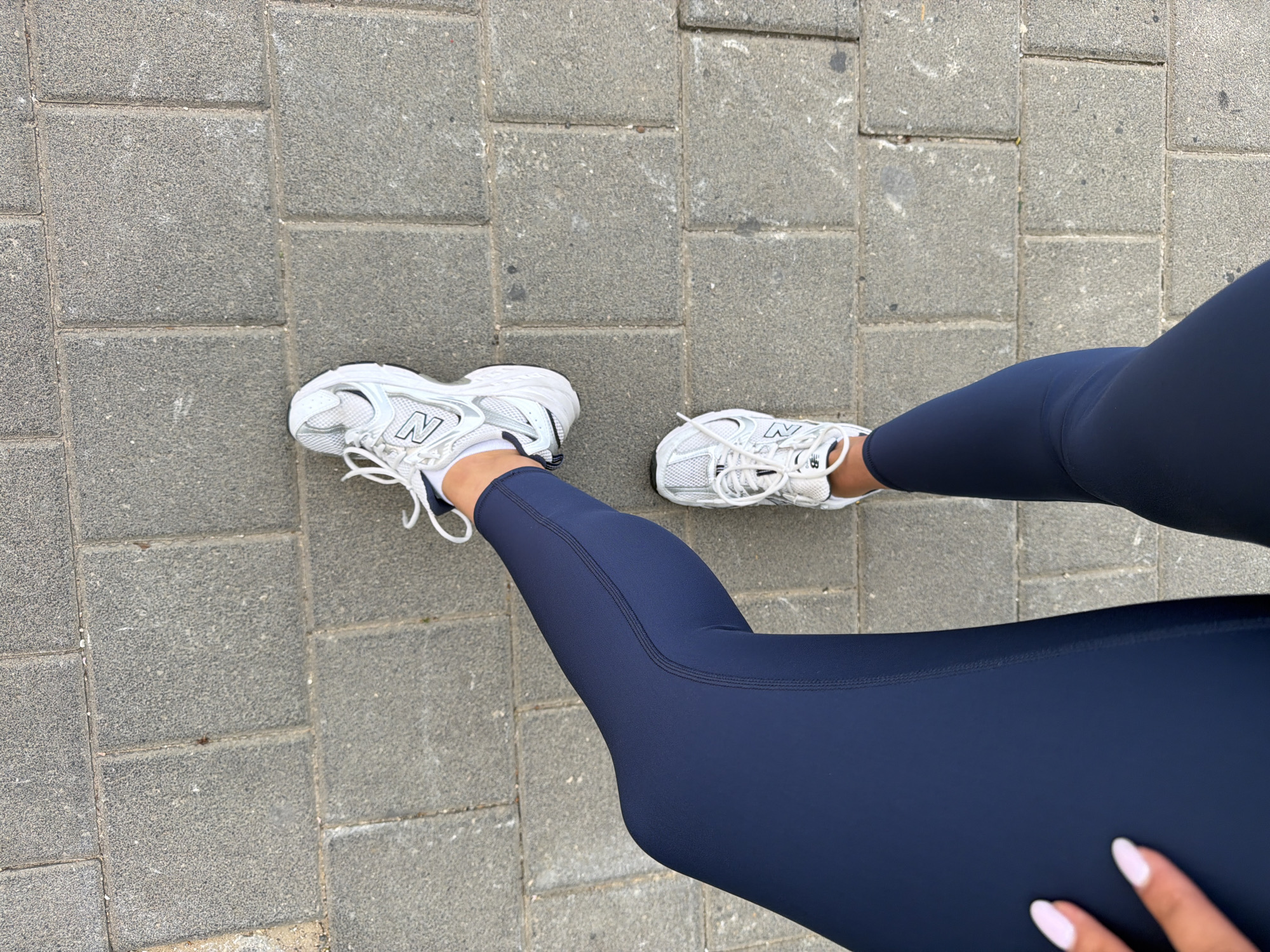 The image presents a top-down view of a person's lower body, specifically their legs and feet, on a paved outdoor surface. The subject is wearing dark blue, form-fitting athletic leggings and white New Balance sneakers. The iconic "N" logo is clearly visible on the side of both shoes. The sneakers feature a design with mesh and synthetic overlays.
The ground beneath the person is a pathway or sidewalk made of grey, rectangular concrete pavers, which have a granular texture. Some subtle variations in color and minor marks are visible on the paving stones. The lighting appears even and diffused, suggesting it is daytime, possibly under an overcast sky, as no harsh shadows are cast.
A hand with light-colored, possibly pink or white, manicured fingernails is visible in the lower right portion of the frame, resting on the person's leg. The pose implies the person is standing or paused during an activity such as walking or exercising. Given the provided context, the scene is set in Tel-Aviv, Israel. The visible attire and setting suggest an active, urban outdoor environment.