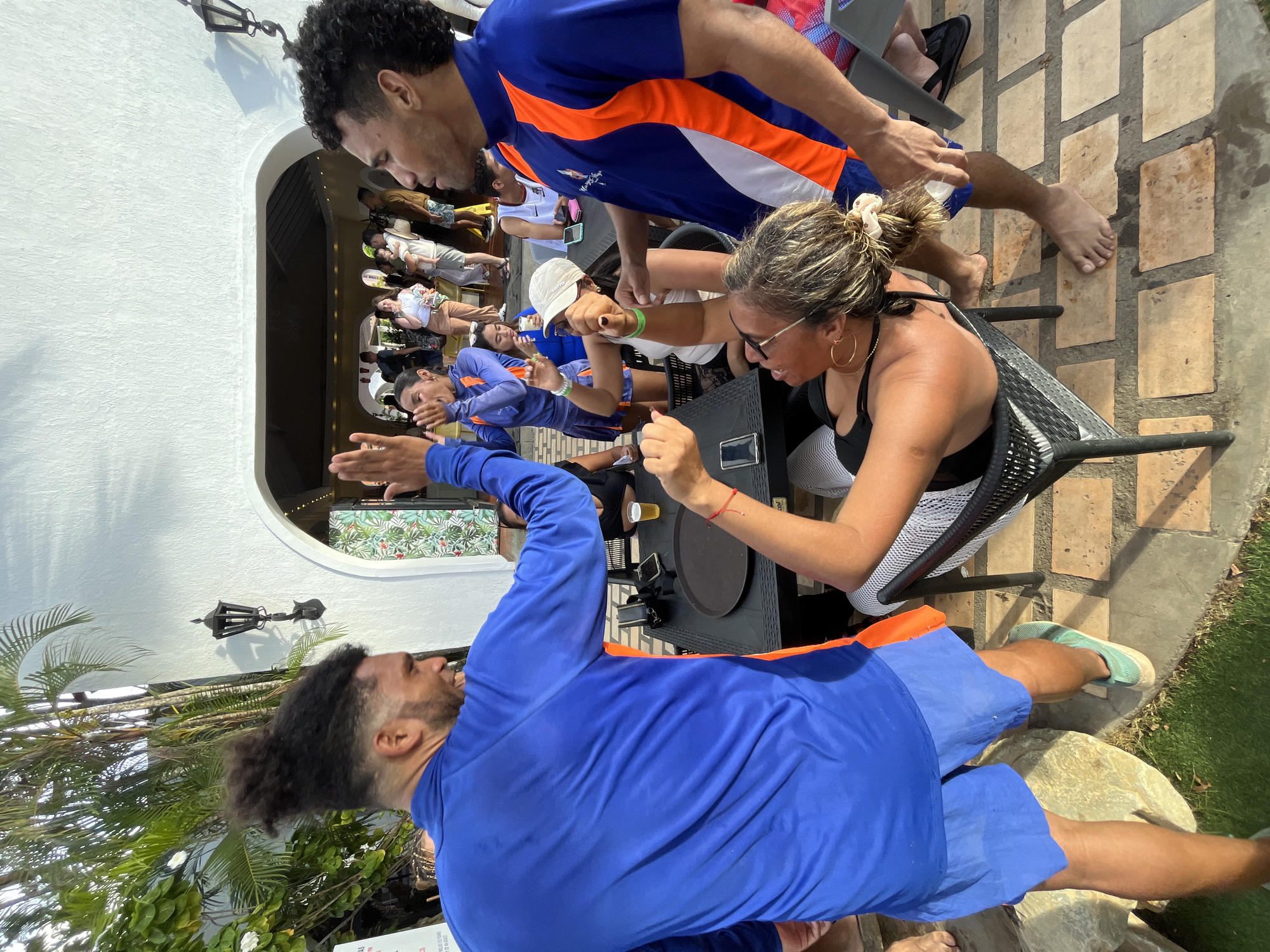The image shows an outdoor scene with people gathered at a restaurant or bar, possibly in Porlamar, Venezuela. Several individuals are visible; two women seated at a table appear to be engaging in a playful arm-wrestling match, with one woman wearing a black top and white shorts, and the other wearing a white top and visor. Two men in blue and orange shirts, possibly teammates, are observing the arm wrestle. Other people can be seen seated at tables in the background, near a large arched entrance. The setting includes tiled flooring, a small patch of grass, and palm trees. A wall with a light fixture is partially visible. Other objects visible include chairs, tables, a beer glass, and a black tray.