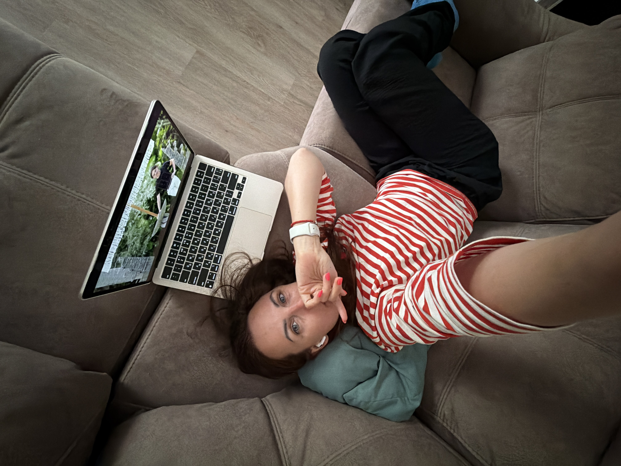A female individual is observed reclining on a light brown upholstered sectional sofa in an indoor domestic setting, identified as Birobidzhan, Russia. She has dark brown hair and blue eyes, and is wearing a red and white horizontally striped long-sleeved shirt and black pants. A teal pillow supports her head. Her left arm is extended, holding the camera to capture a selfie-style image. Her right hand, with pink nail polish, is positioned near her mouth. She wears white wireless earbuds in her right ear, a white smartwatch on her left wrist, and a red string bracelet on her left wrist. A silver-toned laptop with a Cyrillic keyboard is open on the sofa beside her, displaying a video of a male figure standing outdoors amidst green foliage. The flooring visible in the background is a light wood-grain material.